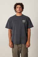 A man with curly hair and a mustache wears the 686 92 Relaxed S/S Tee by 686—a loose, faded black 100% cotton t-shirt with a white logo—paired with patterned gray-green shorts, standing against a plain light background.