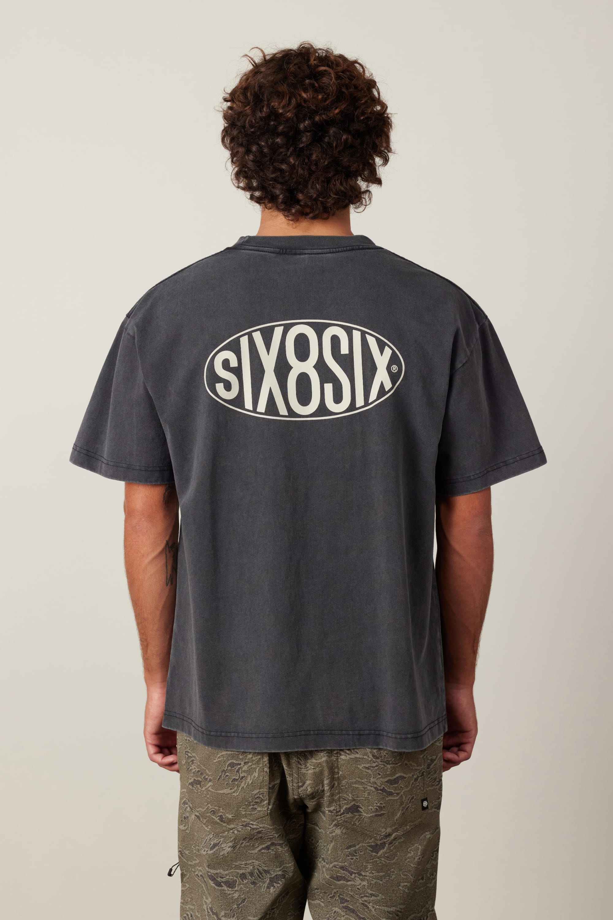 A person with curly hair is seen from behind wearing the 686 92 Relaxed S/S Tee by 686—a faded black, 100% cotton men's t-shirt featuring a large SIX8SIX oval logo on the back—paired with dark-patterned pants against a plain light background.