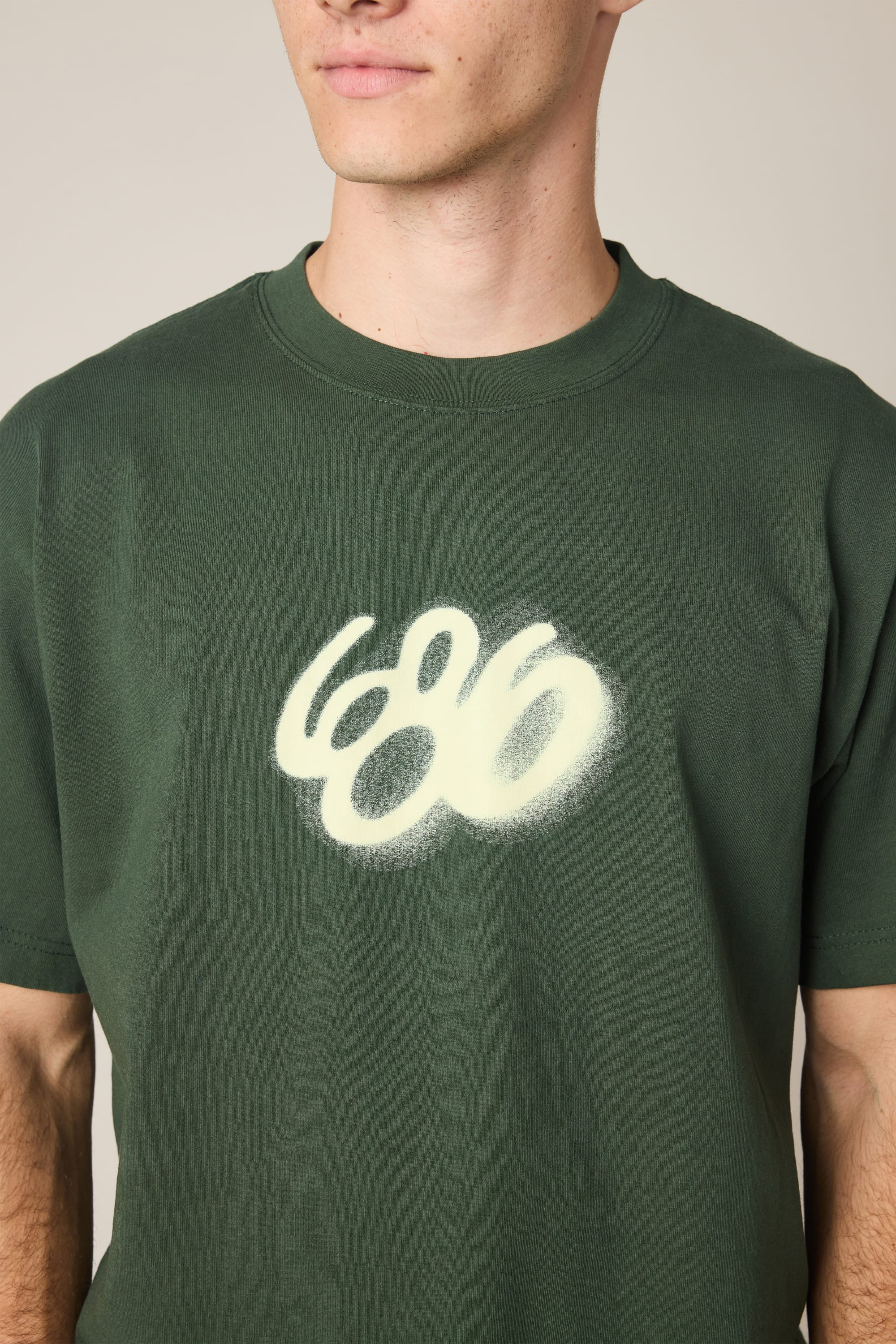 A person wearing the 686 Spray Relaxed SS Tee‚Äîa dark green men‚Äôs t-shirt featuring a white, spray-painted 686 logo. Only the lower face and upper torso are shown, emphasizing its 100% cotton fabric.