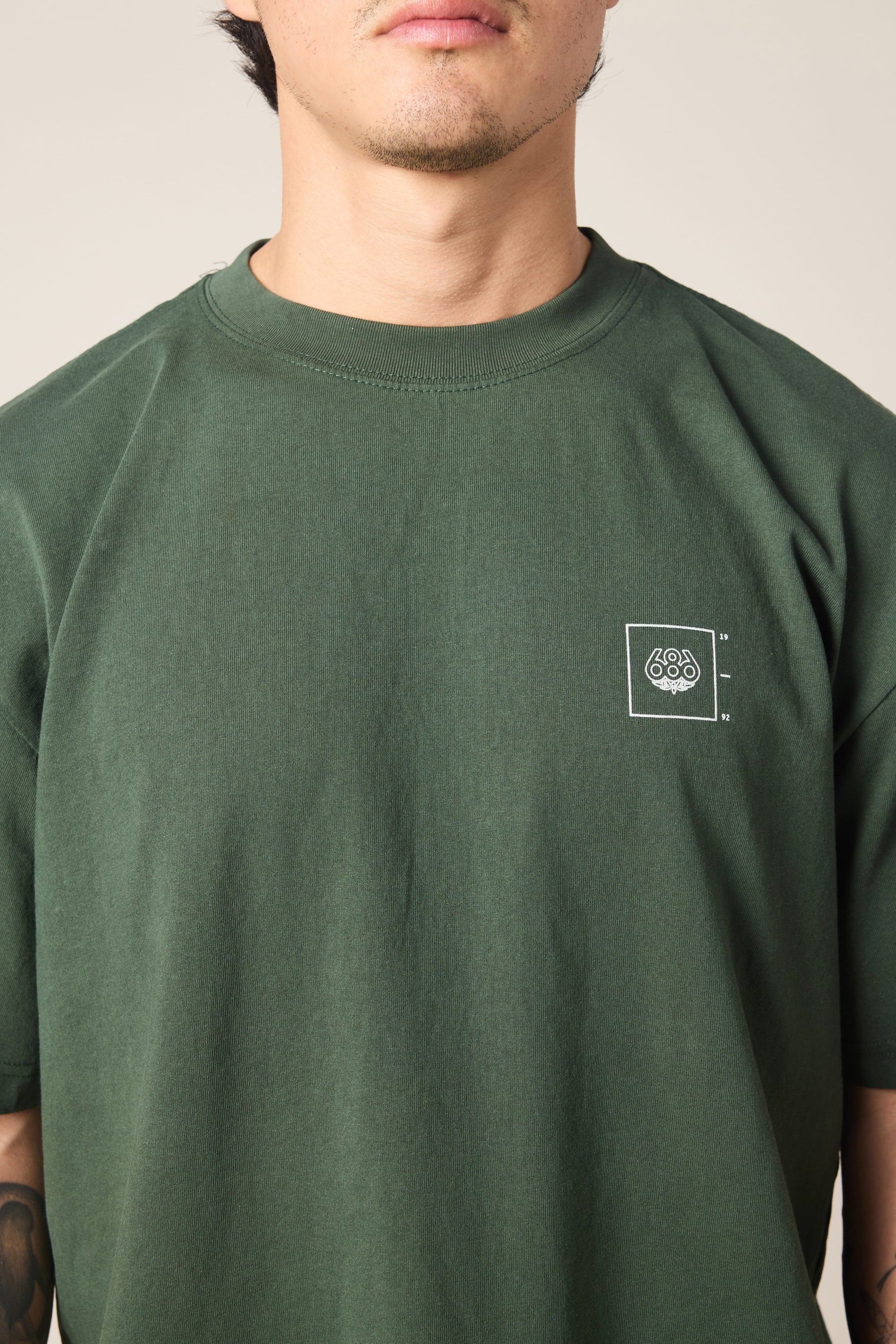 A person wears the 686 Peaks Relaxed SS Tee, a plain dark green men's t-shirt featuring a small white 686 logo on the left chest. Made from soft 100% cotton, it's shown from shoulders to above the mouth against a neutral background.