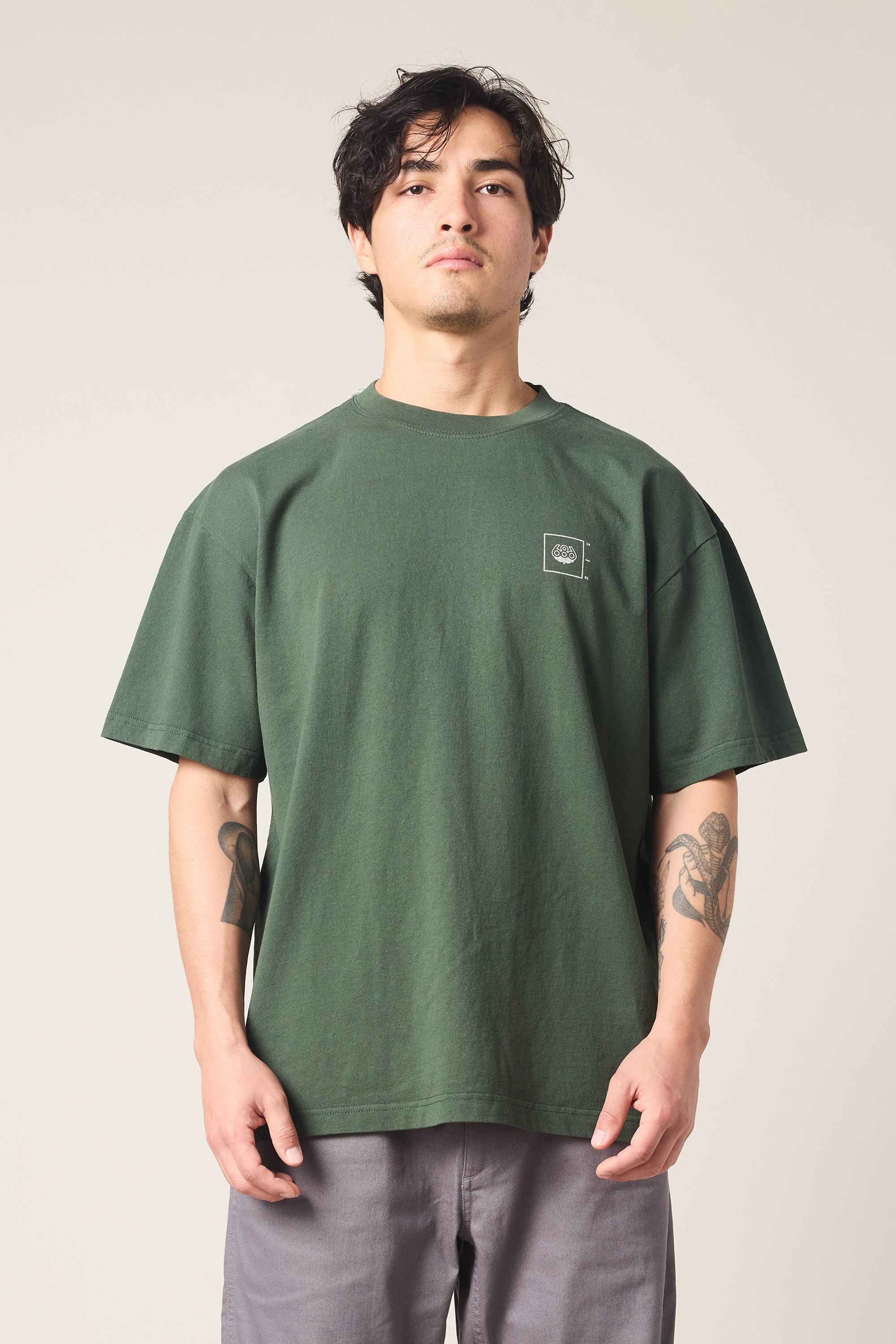A person with dark hair wears the 686 Peaks Relaxed SS Tee by 686 and gray pants, standing forward against a plain light background. Tattoos cover both forearms, and they have a neutral expression.