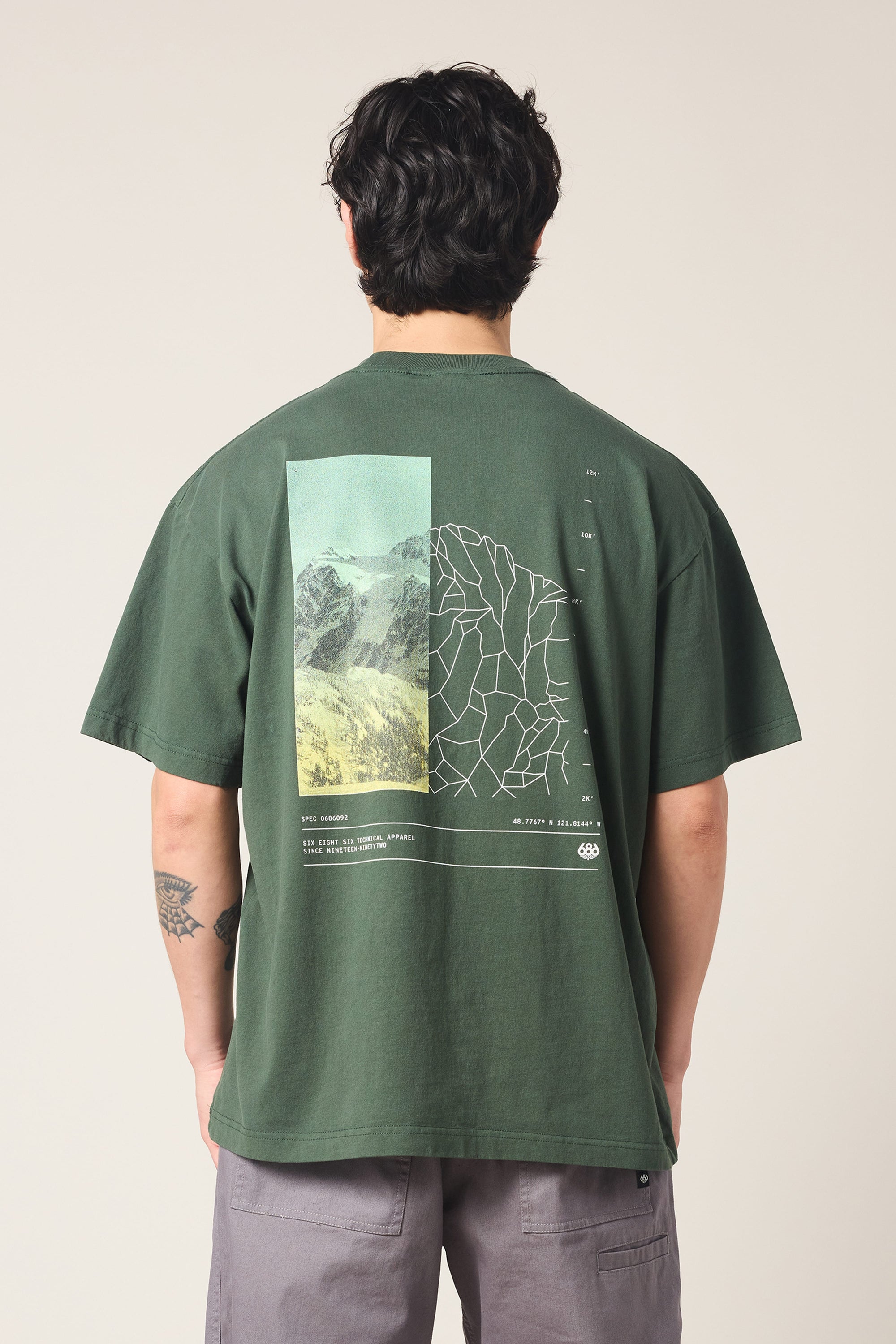 A person with dark hair and a left arm tattoo stands facing away, wearing the 686 Peaks Relaxed SS Tee by 686—a dark green 100% cotton men's t-shirt featuring a mountain and abstract line graphic on the back.