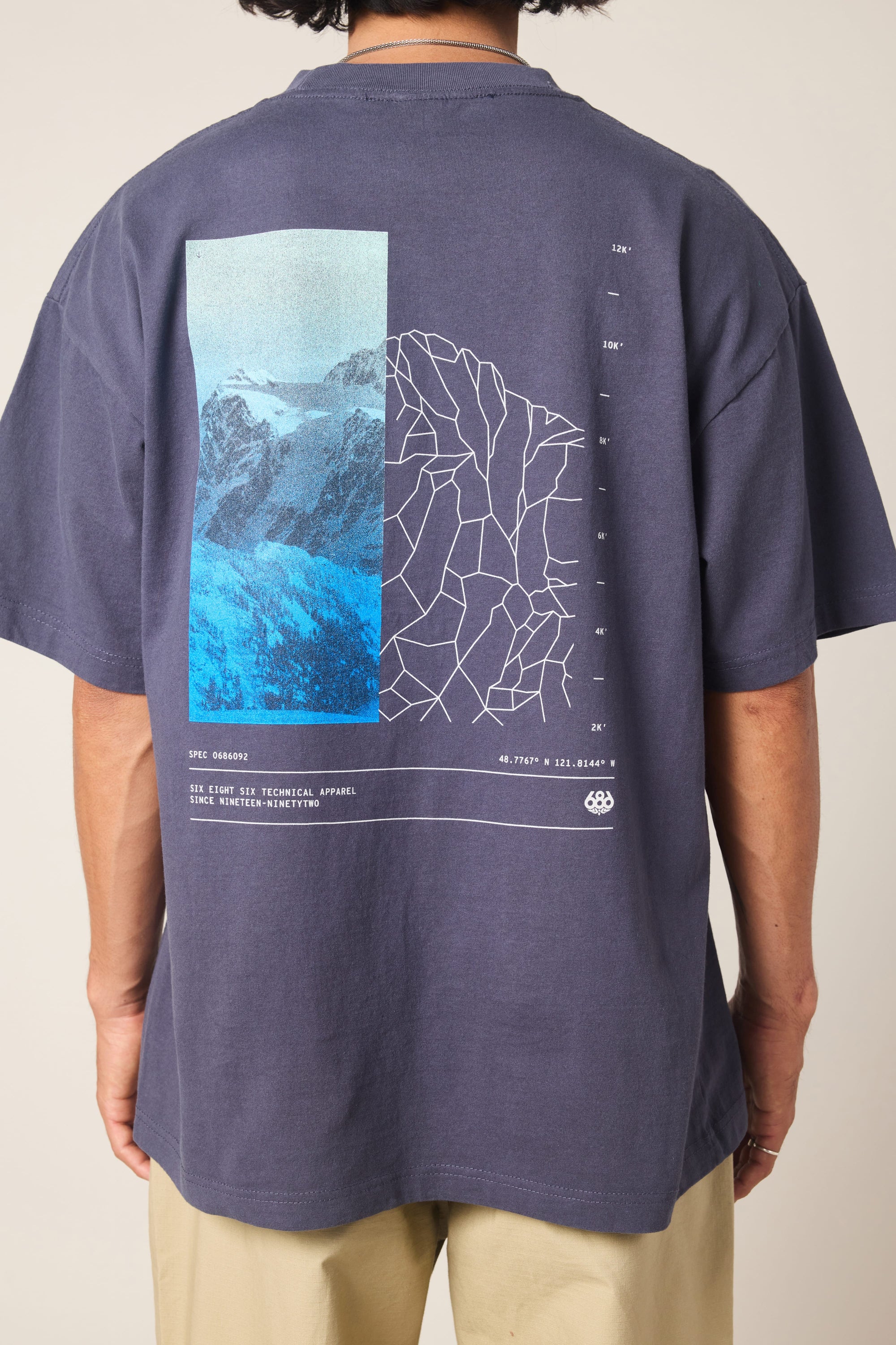 A person is shown from behind wearing the 686 Peaks Relaxed SS Tee by 686, a 100% cotton navy men's t-shirt with a back graphic featuring blue-toned mountains, a white geometric map design, and technical text.