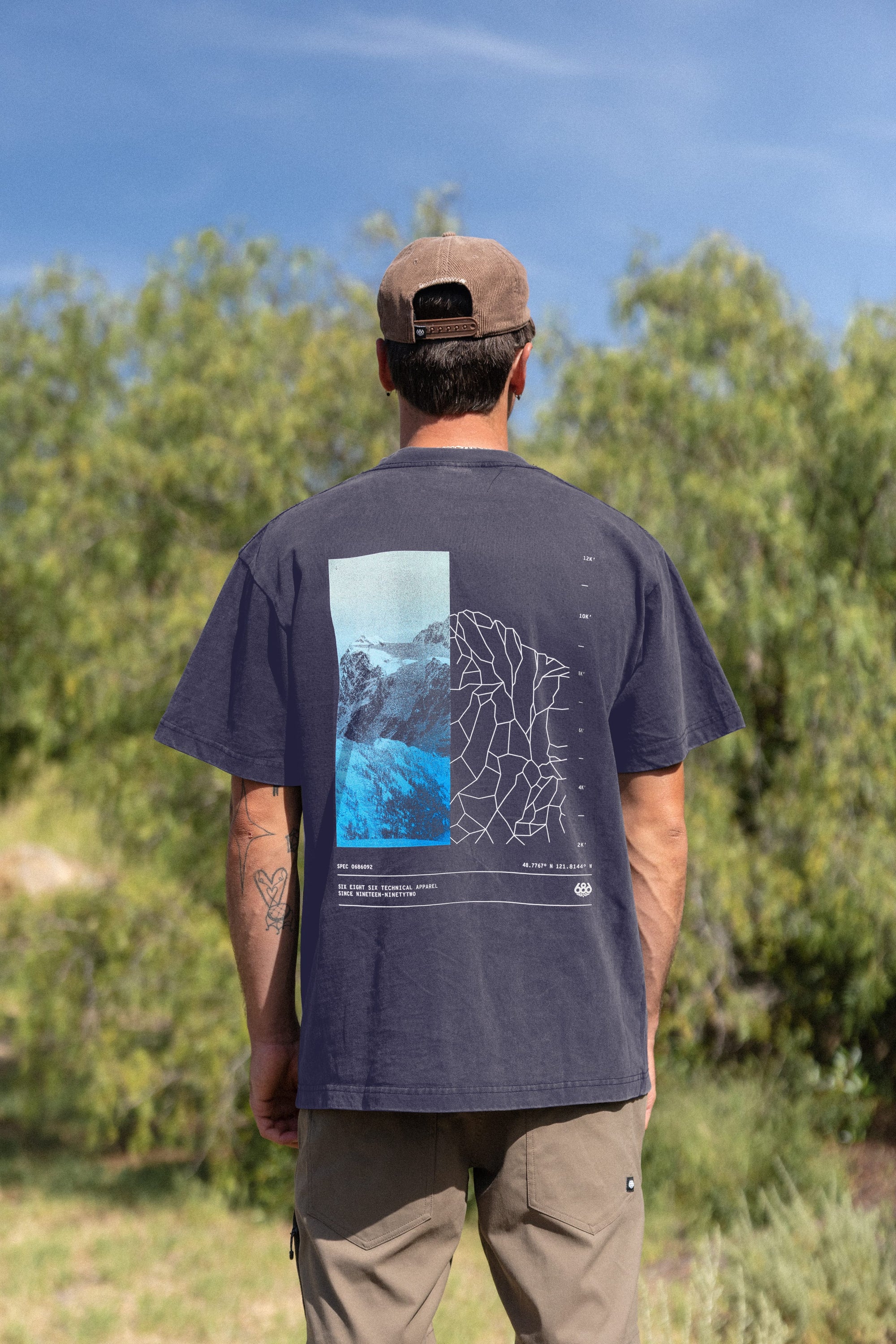 A man stands outdoors among green trees under a blue sky, wearing the 686 Peaks Relaxed SS Tee by 686—a black, 100% cotton tee with abstract graphics and a mountain photo on the back—paired with a brown cap and light khaki pants.