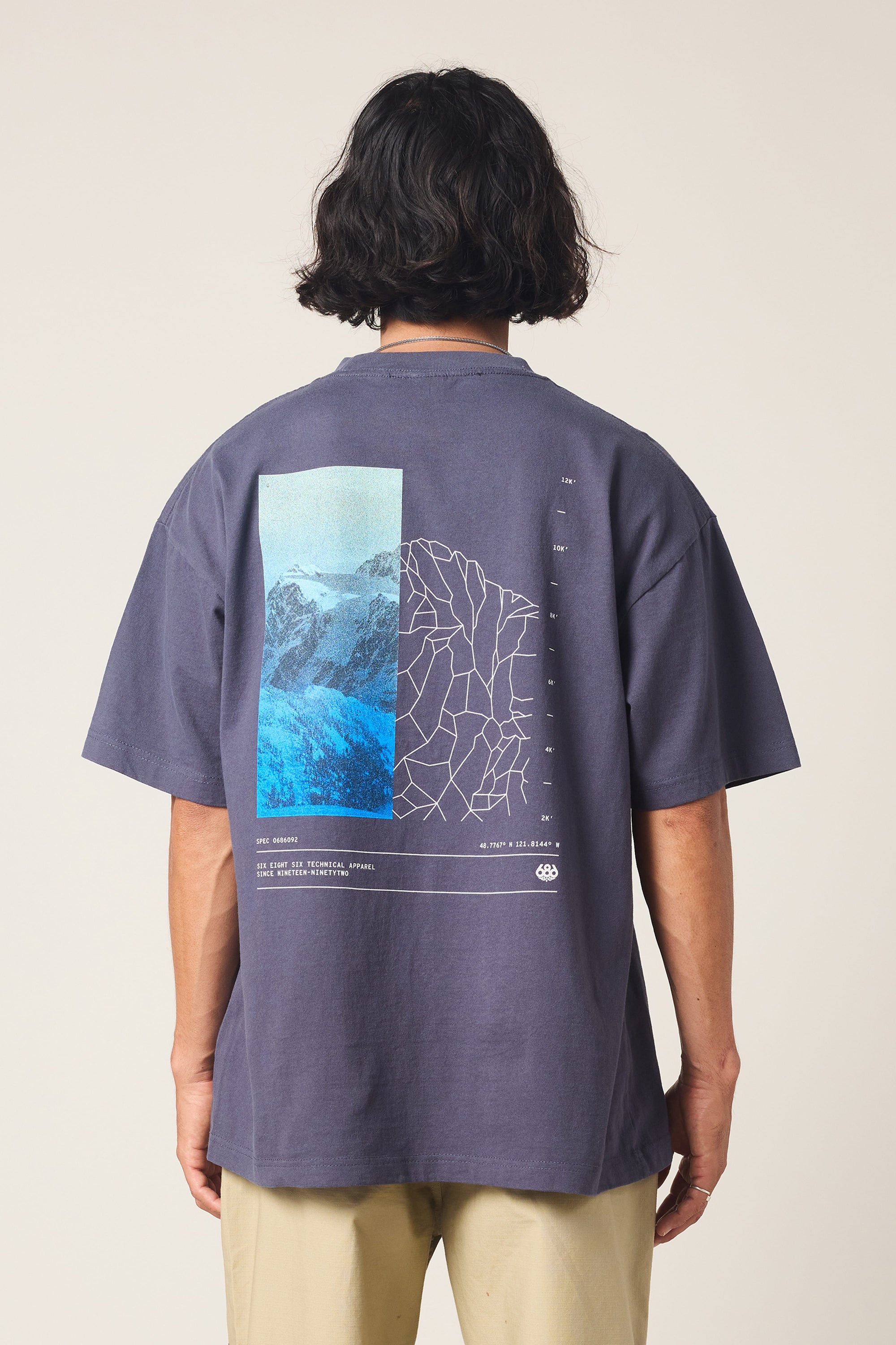 A person with medium-length dark hair, seen from the back, wears the 686 Peaks Relaxed SS Tee by 686—a dark gray 100% cotton men's shirt featuring a blue mountain and white contour map graphic—with beige pants.