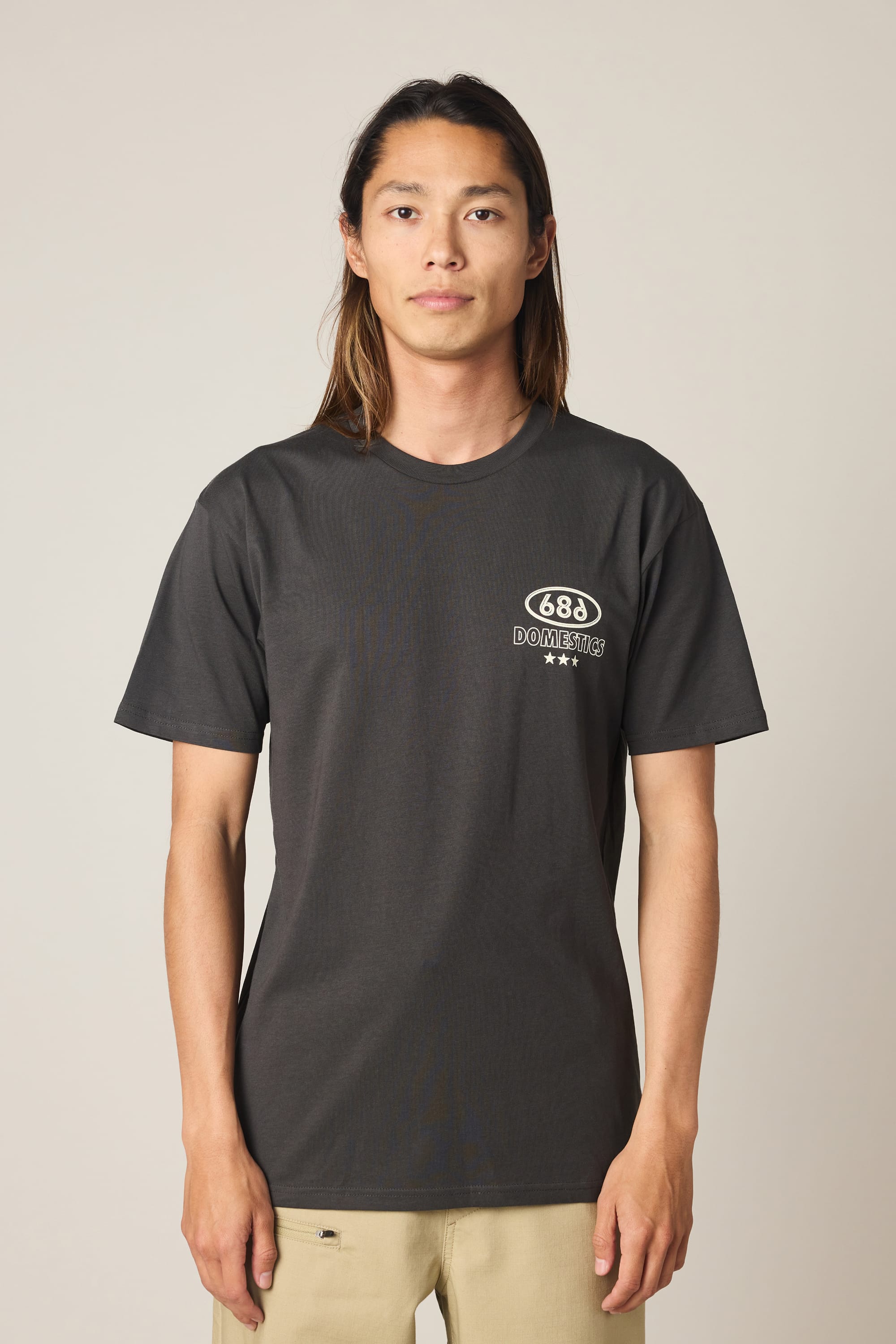 A person with long hair is wearing the 686 Men's Domestics Authentic SS Tee, a black 100% cotton t-shirt by 686 with a small white Domestics logo on the left chest, standing against a plain light background.