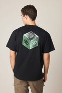 A person with short brown hair, seen from behind, wears the 686 Men's Unit Relaxed SS Tee—a black 100% cotton T-shirt by 686 featuring a green and white cube graphic with skull designs on the back. He is also wearing brown pants.