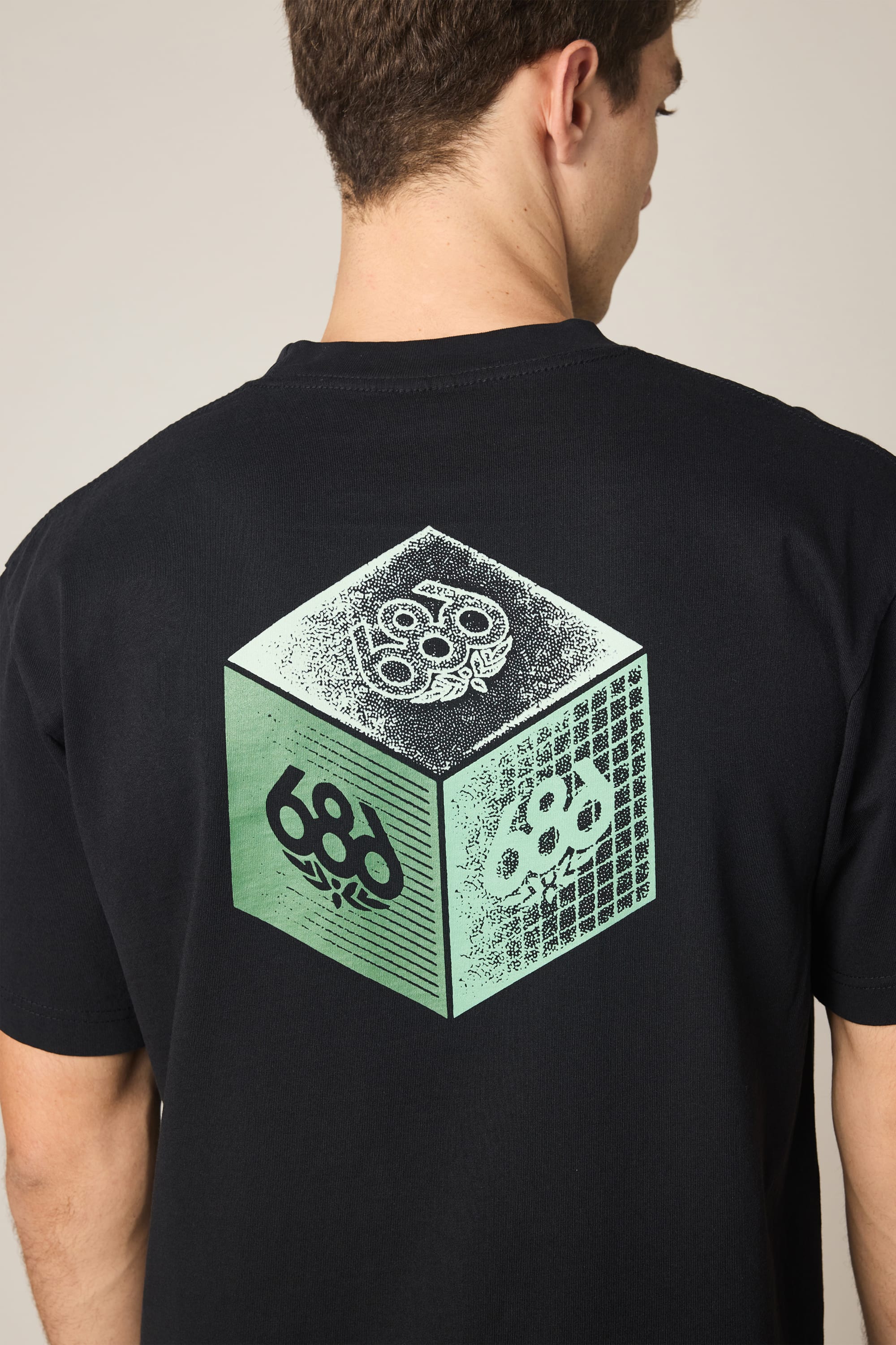 A person wears the 686 Men's Unit Relaxed SS Tee, a black 100% cotton shirt with a bold cube graphic, "686," and green and white patterns on the back. The shirt is shown from behind, the wearer’s head turned slightly to the side.