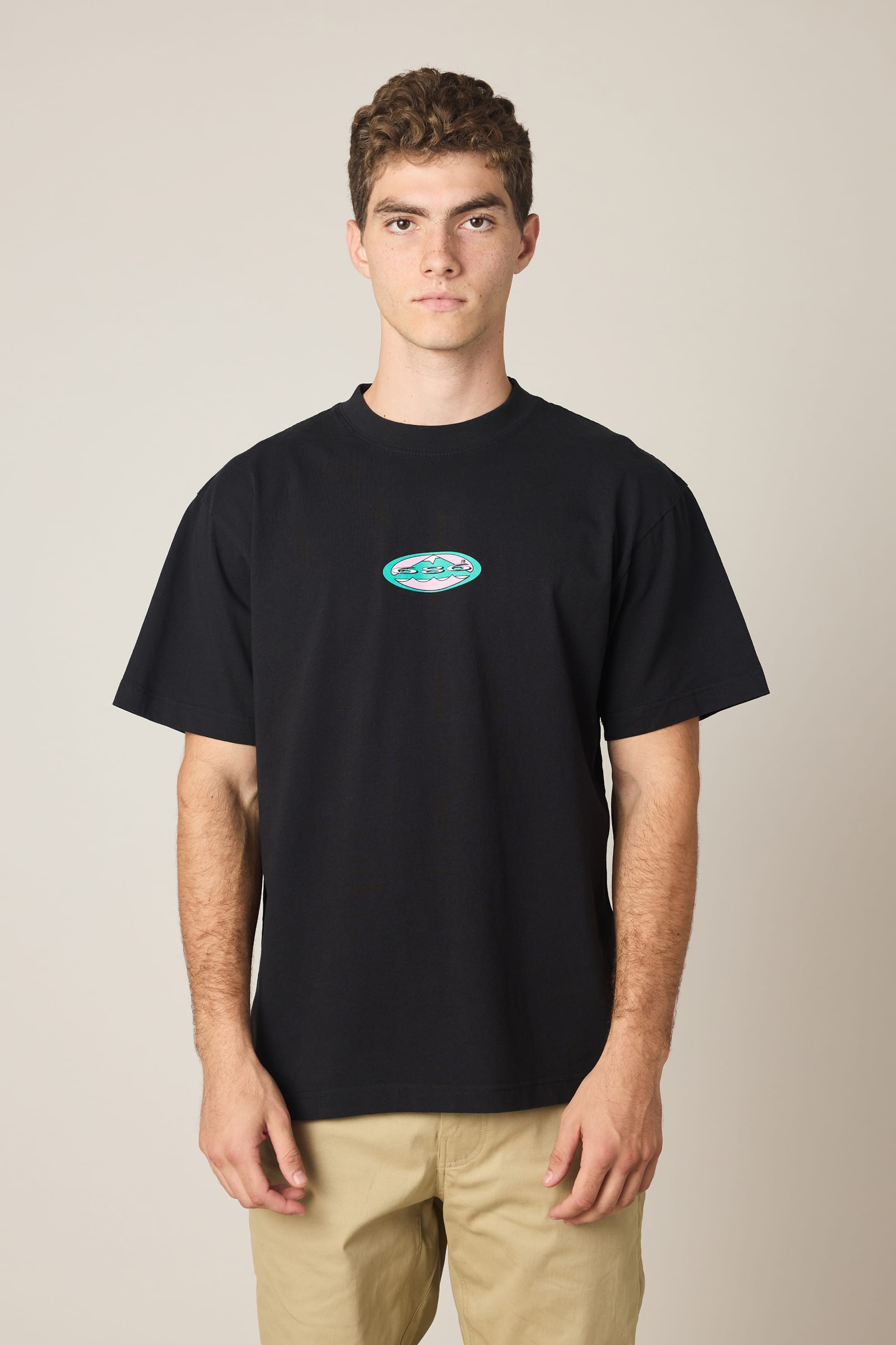A young man with short curly hair wears the 686 Men's Jib Relaxed SS Tee by 686, featuring a small colorful oval chest logo and beige pants, standing against a plain light background and looking straight at the camera.