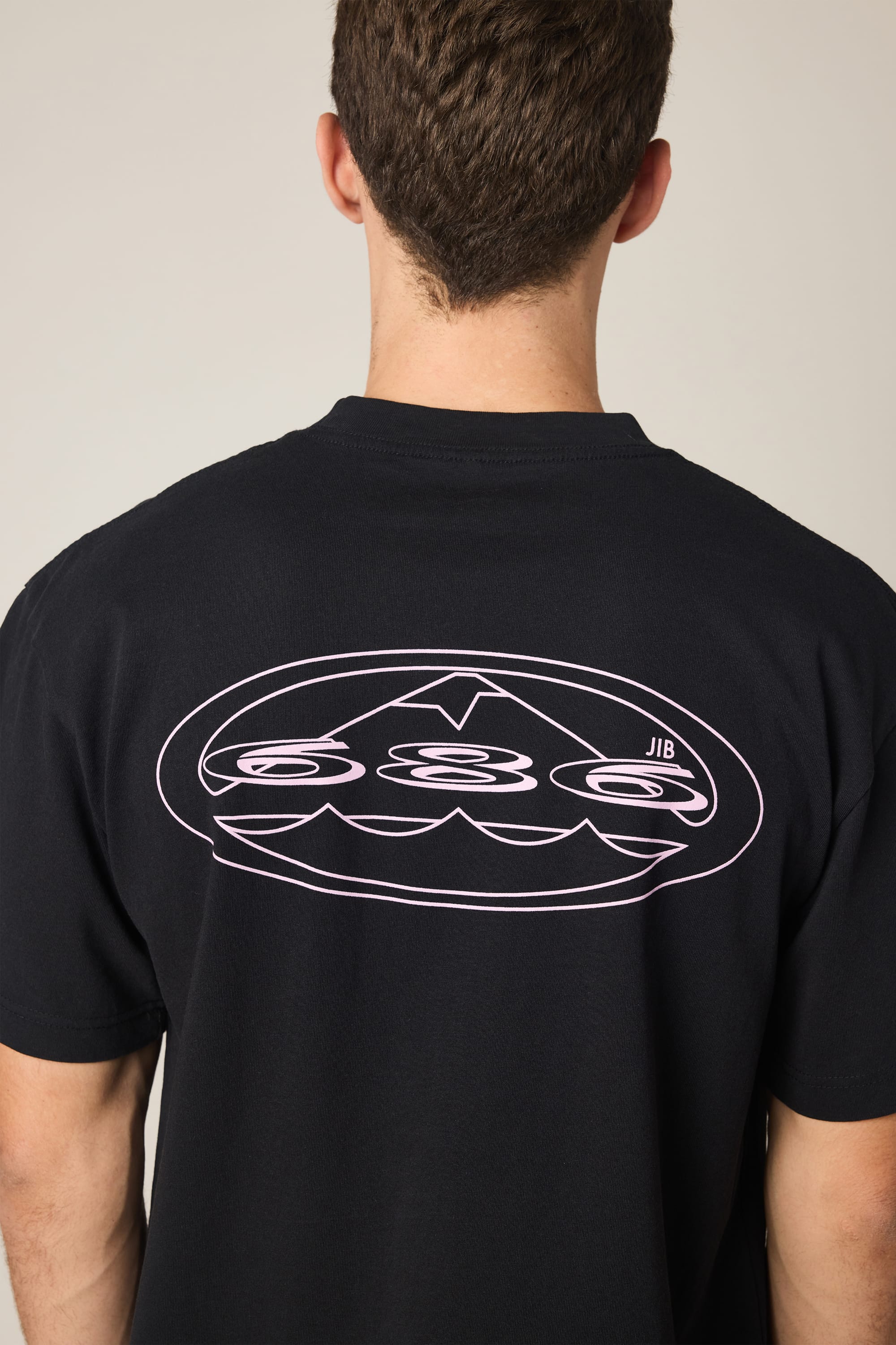 A person with short brown hair is seen from behind wearing the 686 Men's Jib Relaxed SS Tee, which features a large white outlined “JIB” graphic with stylized waves and an arrow within an oval on the back.