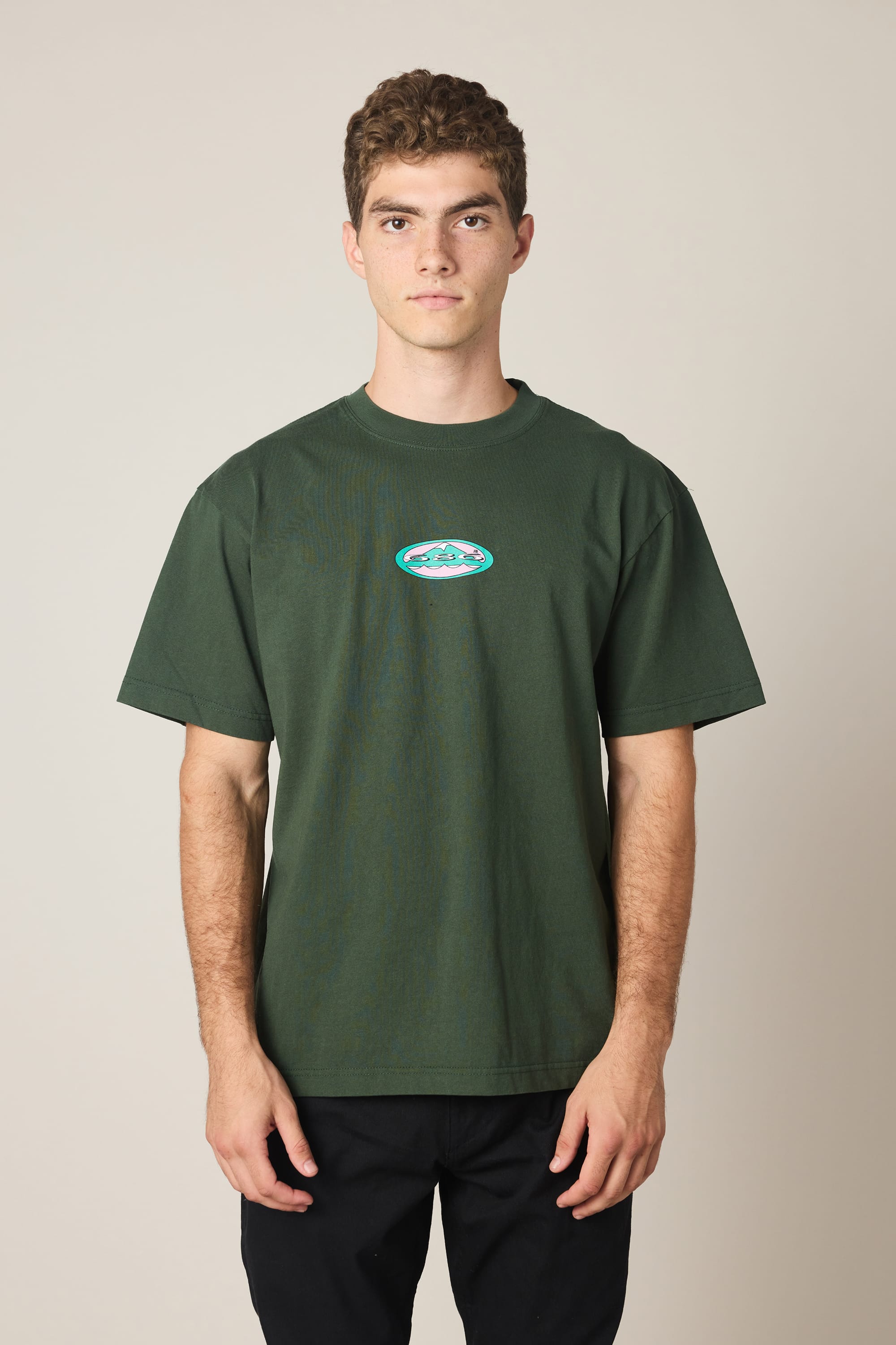 A young man with short curly hair wears the 686 Men's Jib Relaxed SS Tee, featuring a small, colorful oval logo on the chest, against a plain beige background. This stylish cotton t-shirt from 686 offers everyday comfort and effortless style.