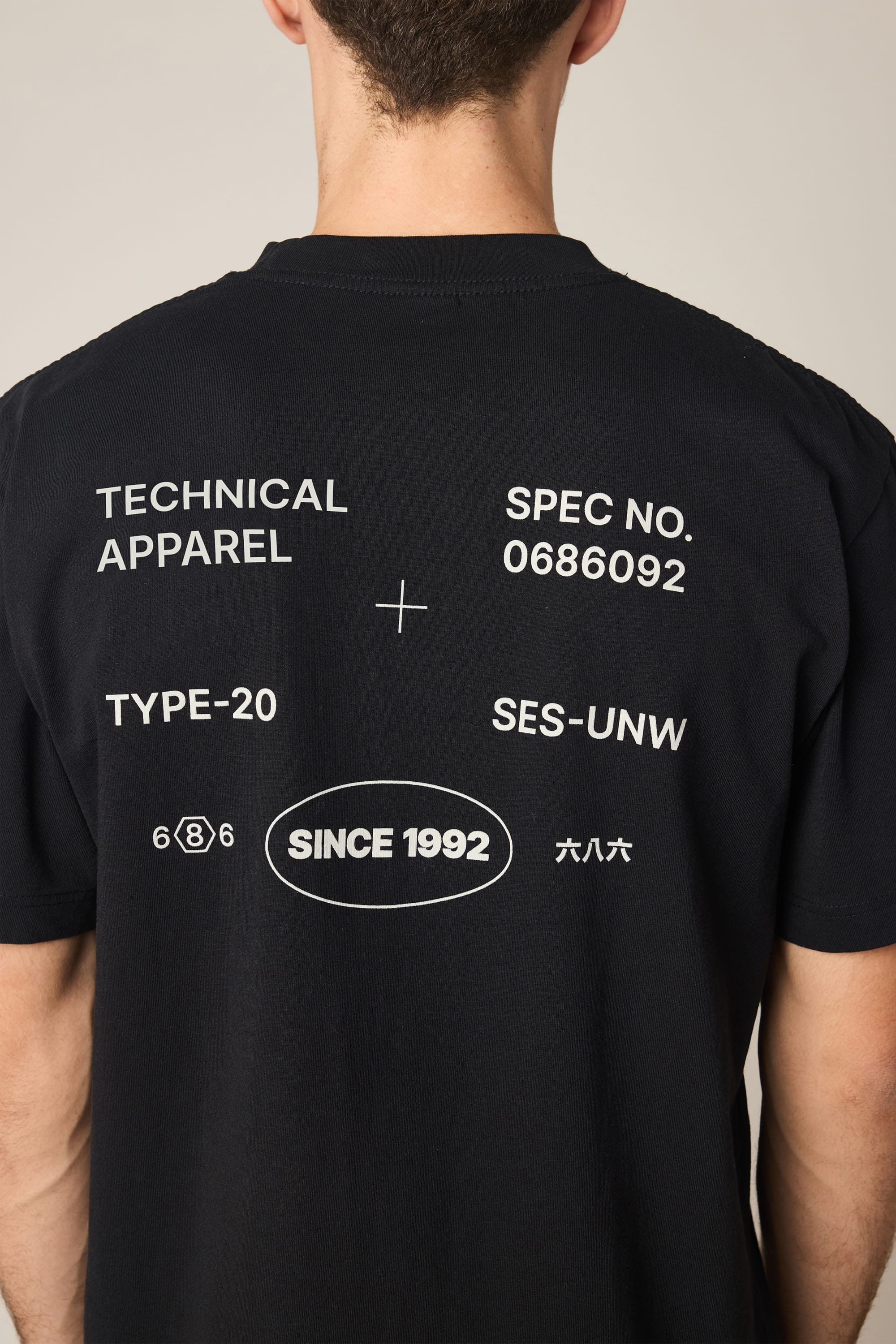 A person is seen from the back wearing the 686 Men's Unwork Spec Relaxed SS Tee by 686, a black 100% cotton t-shirt with white text and symbols: TECHNICAL APPAREL, SPEC NO. 0686092, TYPE-20, SES-UNW, SINCE 1992, numbers, and Chinese characters.