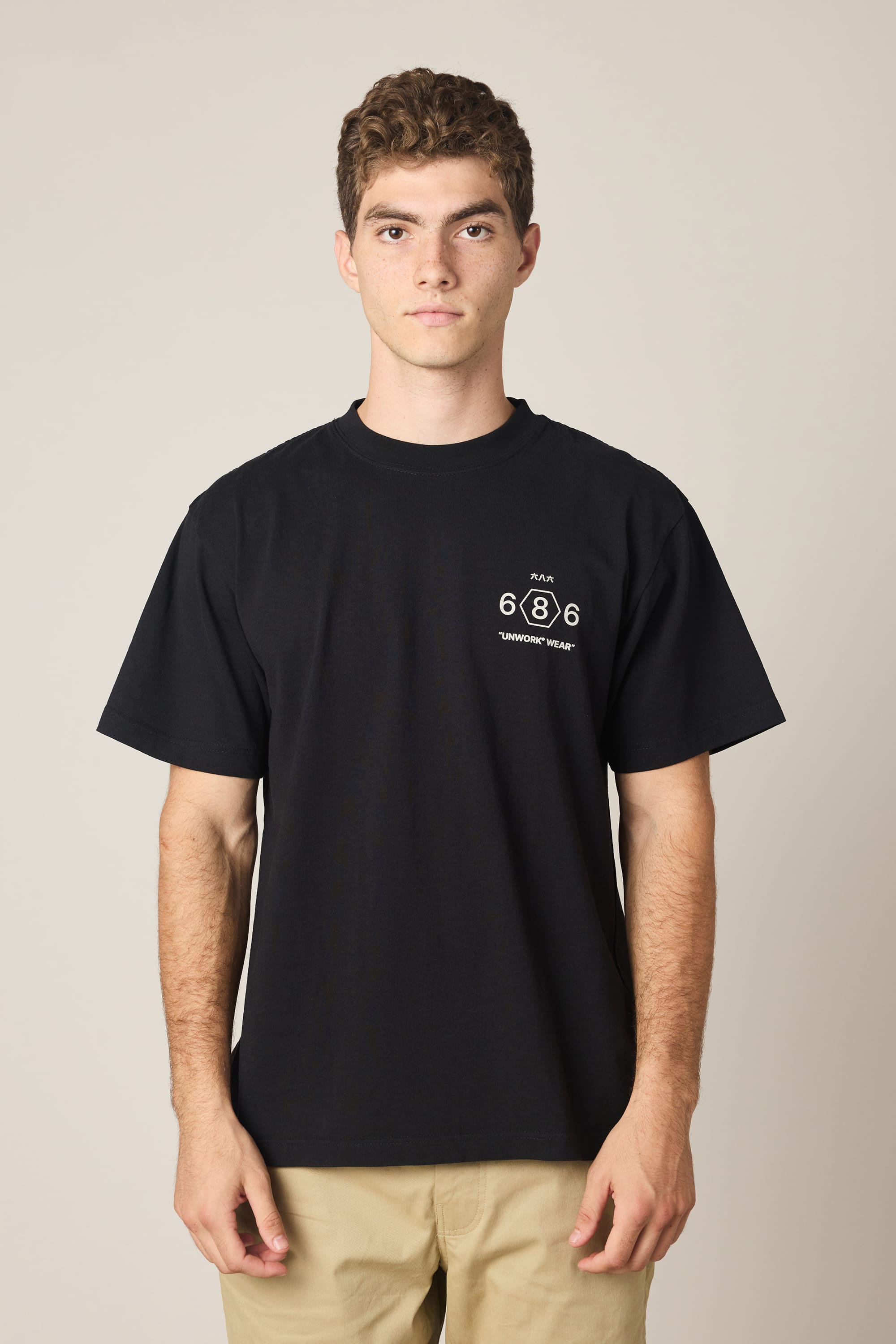 A young man with short curly hair wears the 686 Men's Unwork Spec Relaxed SS Tee, a black 100% cotton shirt with a small white 686 logo and "ANYWHERE WEAR" text on the chest, paired with beige pants against a plain light background.