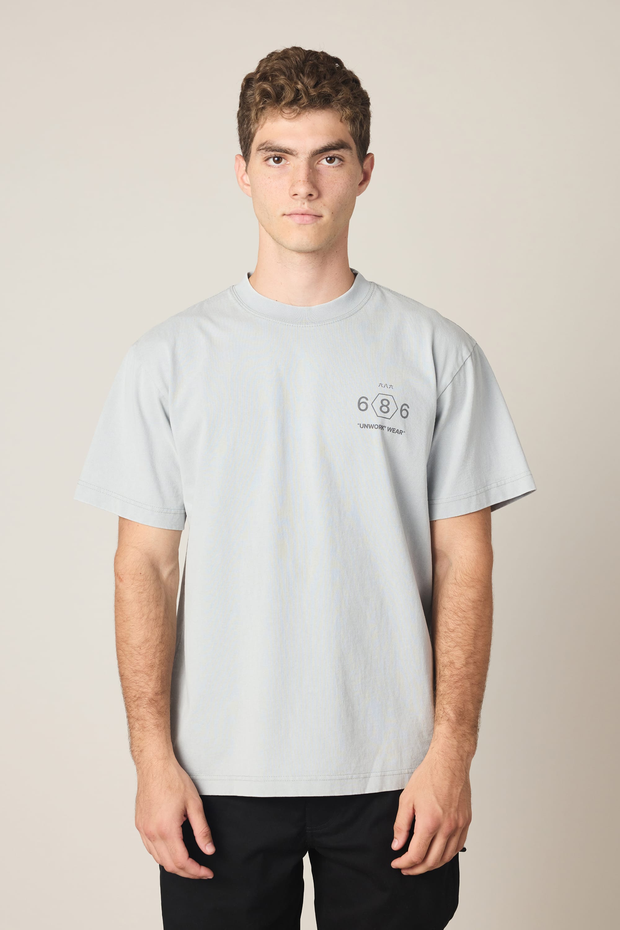 A young man with short curly hair wears the 686 Men's Unwork Spec Relaxed SS Tee, a light gray 100% cotton t-shirt from 686 featuring "686" and small text on the left chest. He stands facing forward against a plain beige background with a neutral expression.