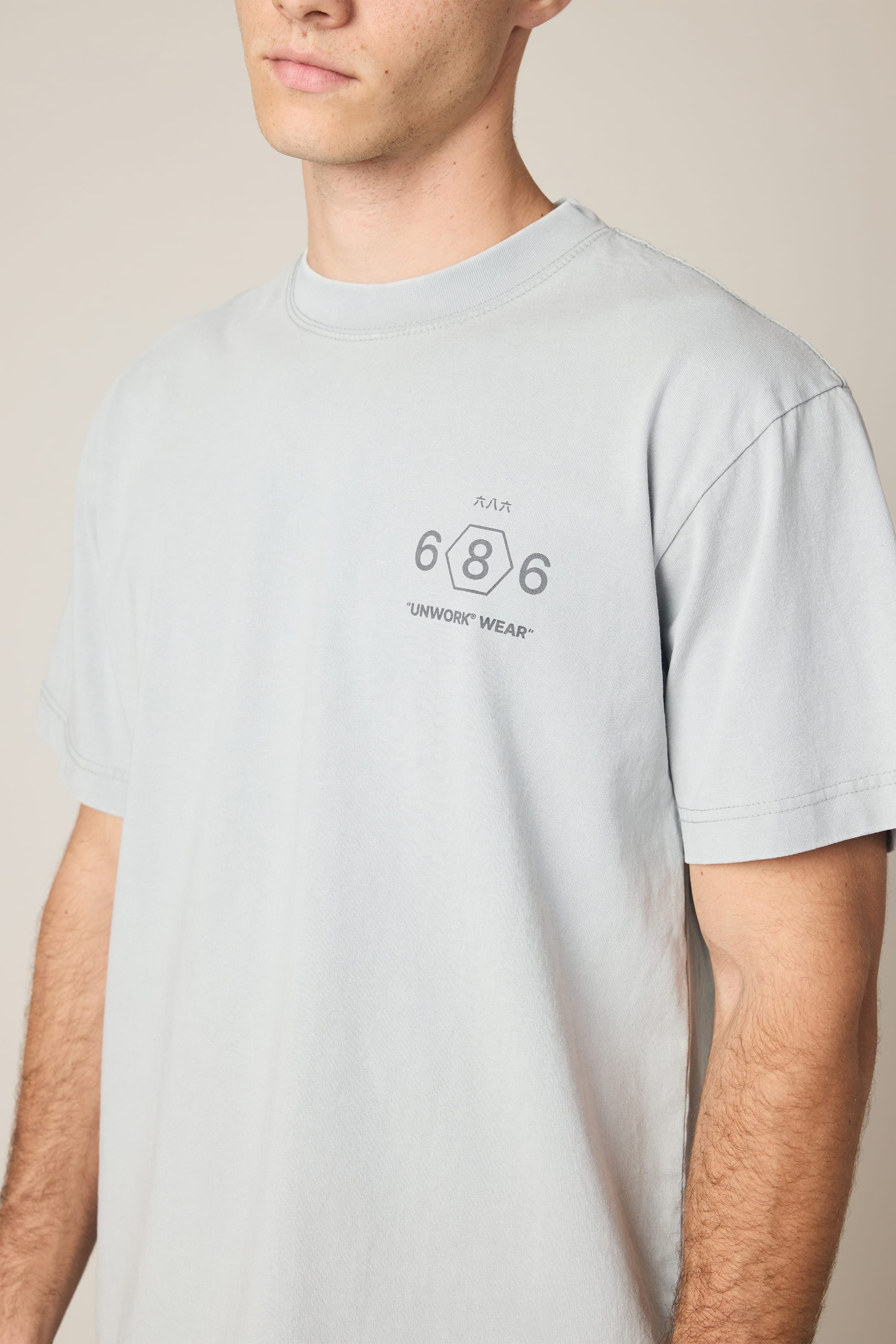 A person wears the 686 Men's Unwork Spec Relaxed SS Tee, a light gray 100% cotton t-shirt with small dark "686" and "UNWORK WEAR" prints on the left chest. Their face is partially out of frame.