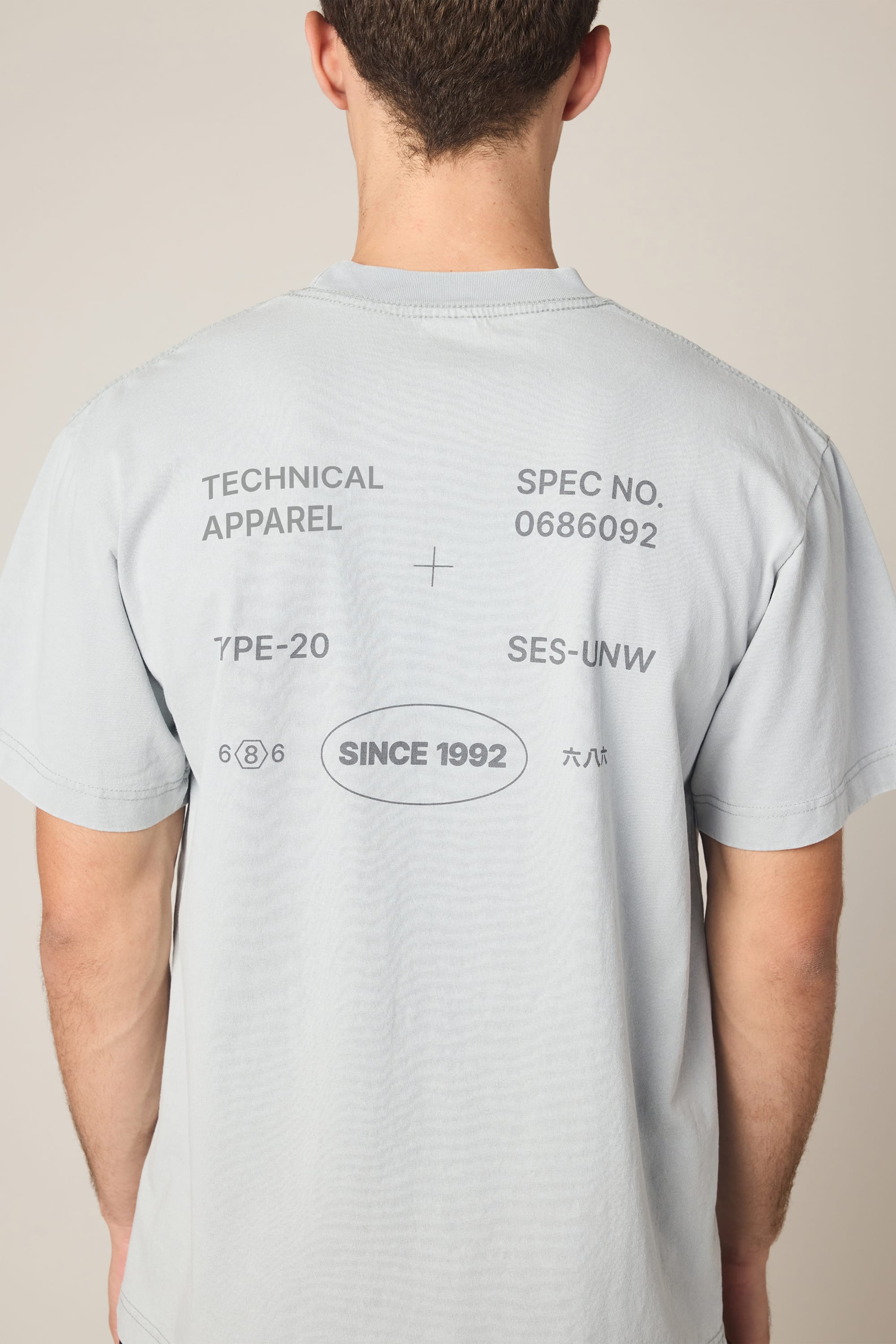A person is seen from the back wearing the 686 Men's Unwork Spec Relaxed SS Tee in light gray, featuring black printed text such as TECHNICAL APPAREL, SPEC NO. 0686092, TYPE-20, SES-UNV, and SINCE 1992 on soft 100% cotton.