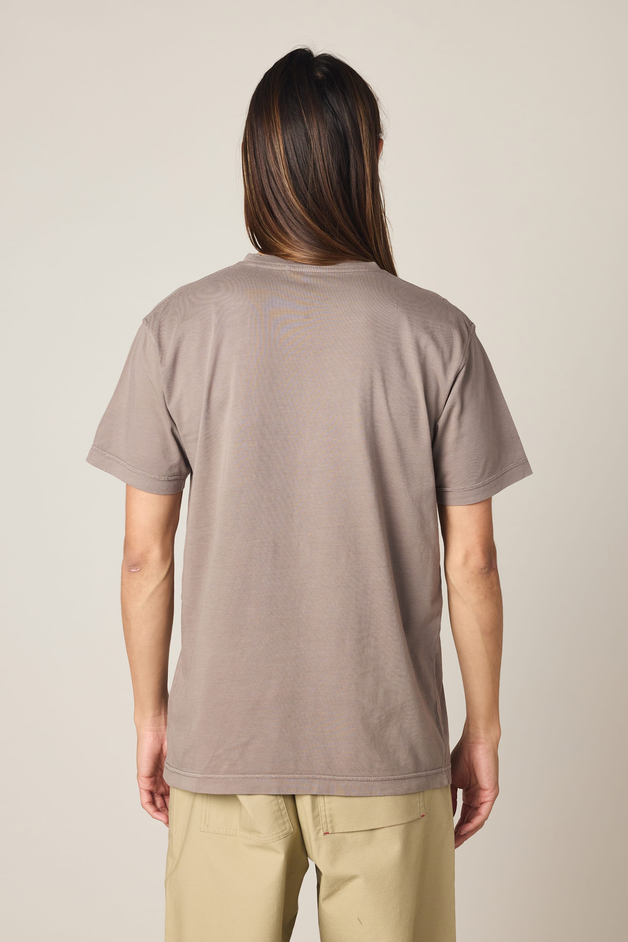 A person with long brown hair stands facing away, wearing the 686 Men's Unwork Authentic SS Pocket Tee by 686 in taupe and beige pants against a plain light background.