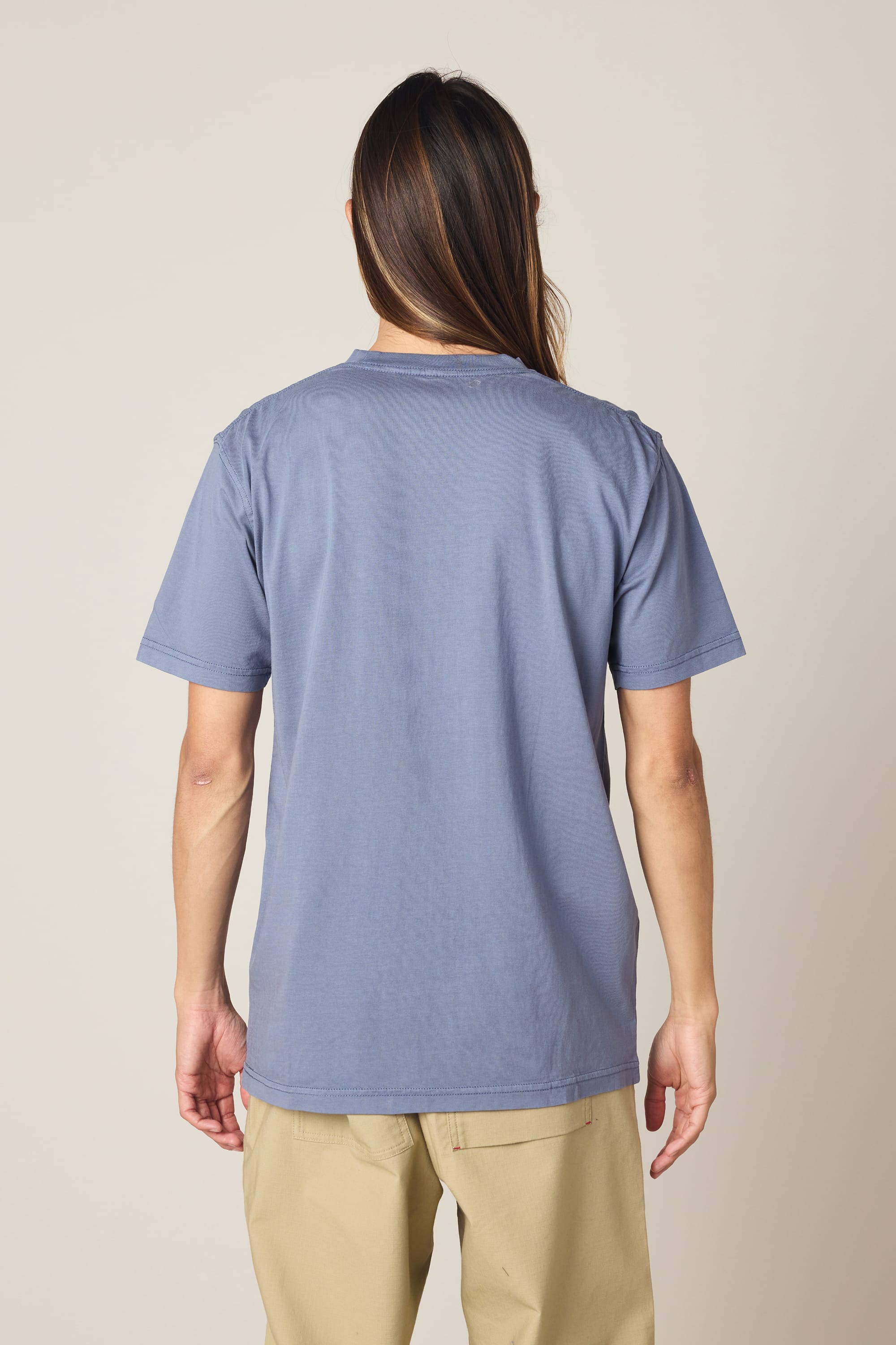 A person with long brown hair is seen from behind, wearing the 686 Men's Unwork Authentic SS Pocket Tee by 686 in blue, along with beige pants, standing against a plain background.
