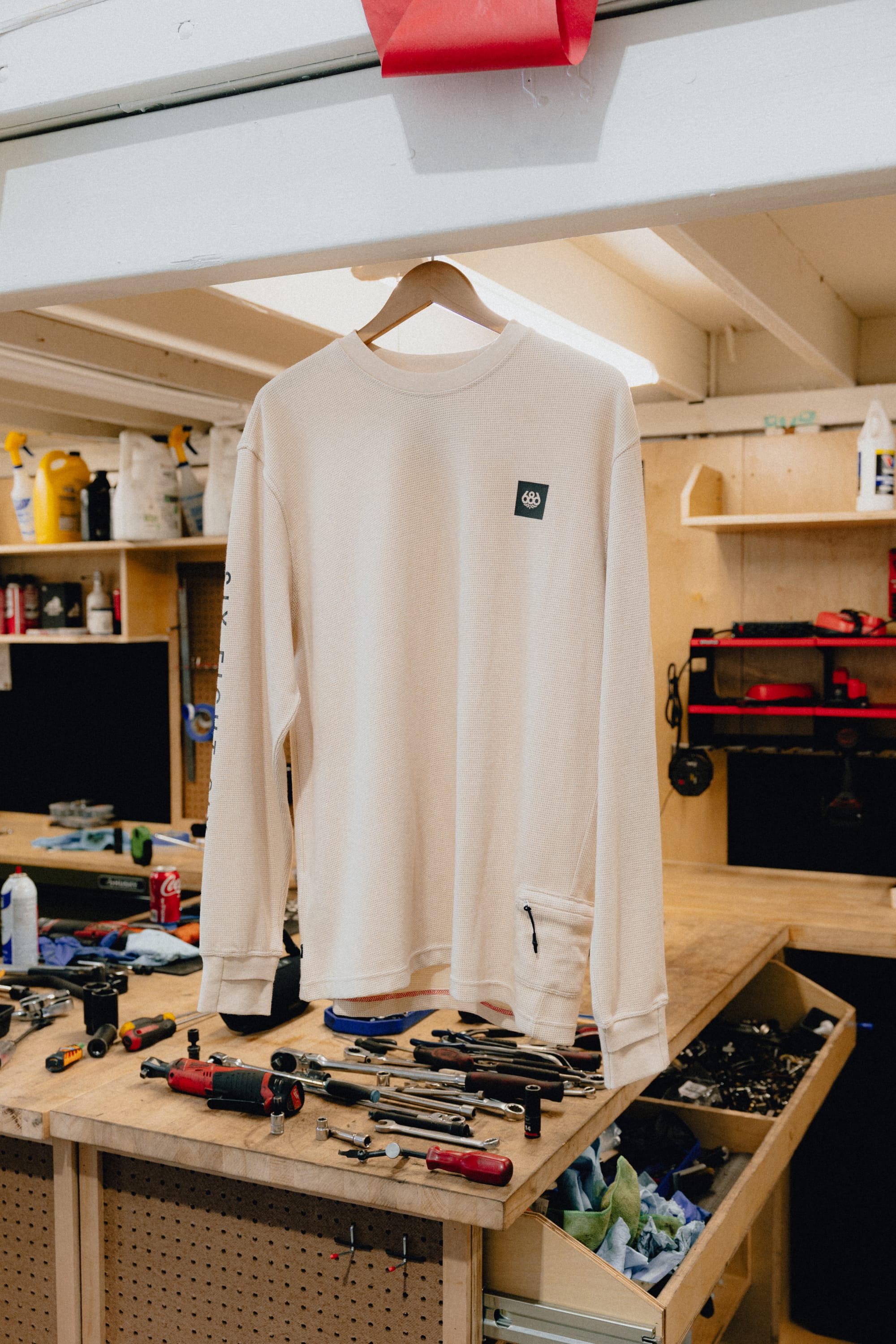 A 686 Men's Let's Chill Waffle LS Crew long sleeve polyester t-shirt in cream hangs on a wooden hanger above a workbench with various hand tools and supplies in a workshop setting.