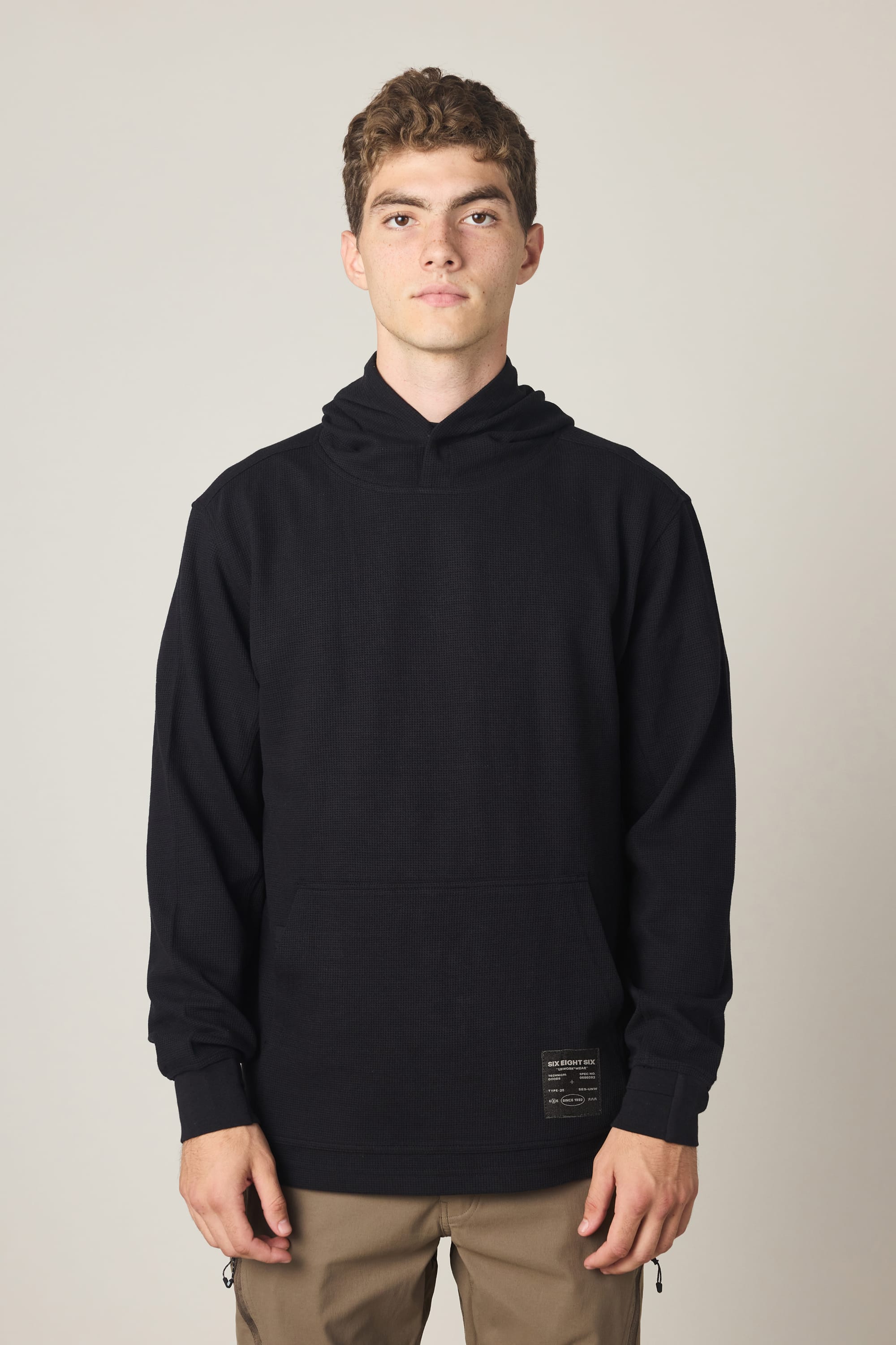 A young man with short curly hair, wearing a 686 Men's Let's Chill Waffle Hoody in black and khaki pants, stands facing forward against a plain light background.