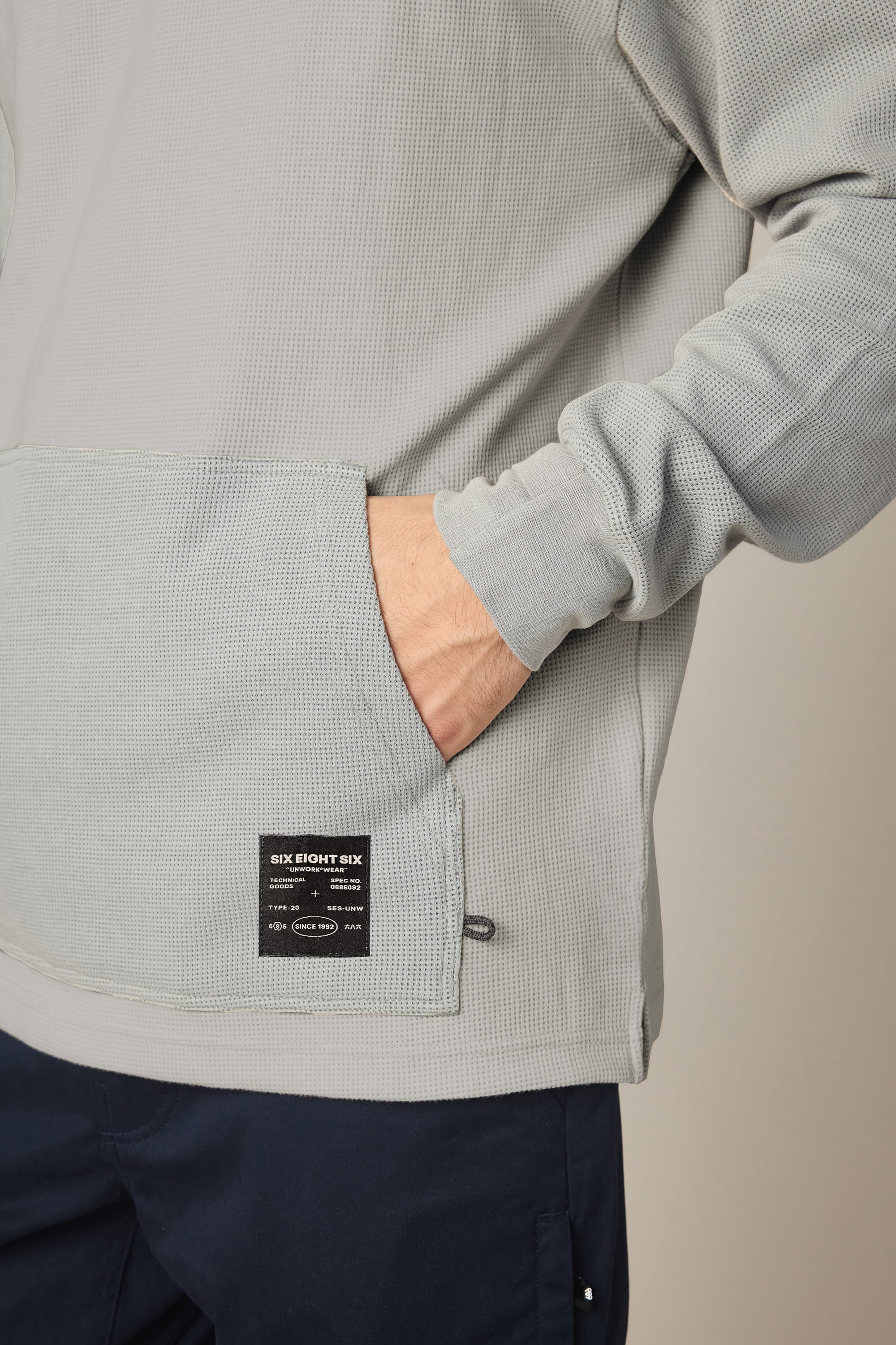 Someone wears the 686 Men's Let's Chill Waffle Hoody—a light grey textured polyester top with one hand in the front pocket, paired with dark pants. A black rectangular label is visible on the pocket.