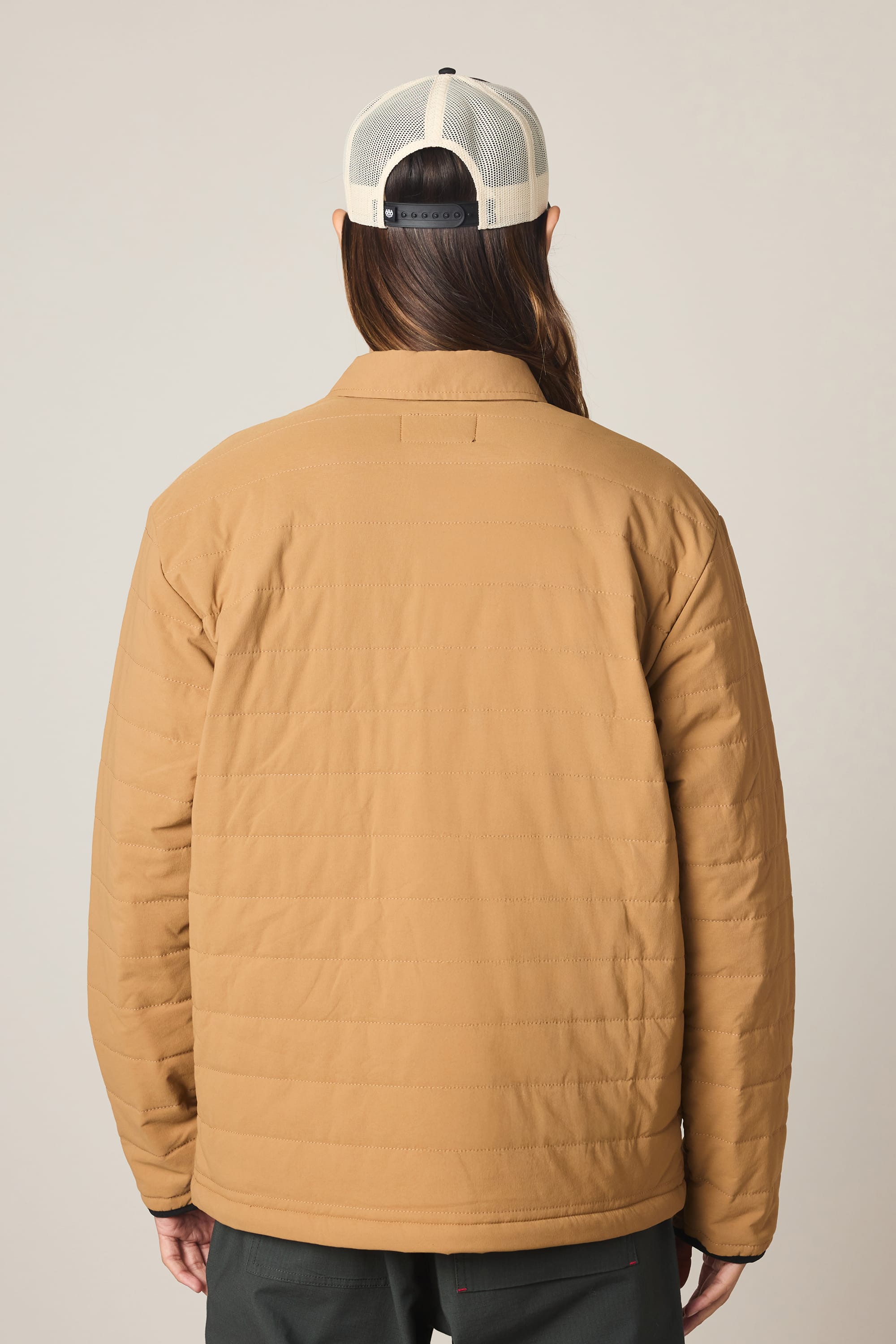 A person wearing the 686 Men's Thermaldry Insulated Shacket in tan, dark pants, and a white mesh baseball cap stands with their back to the camera against a plain light background.