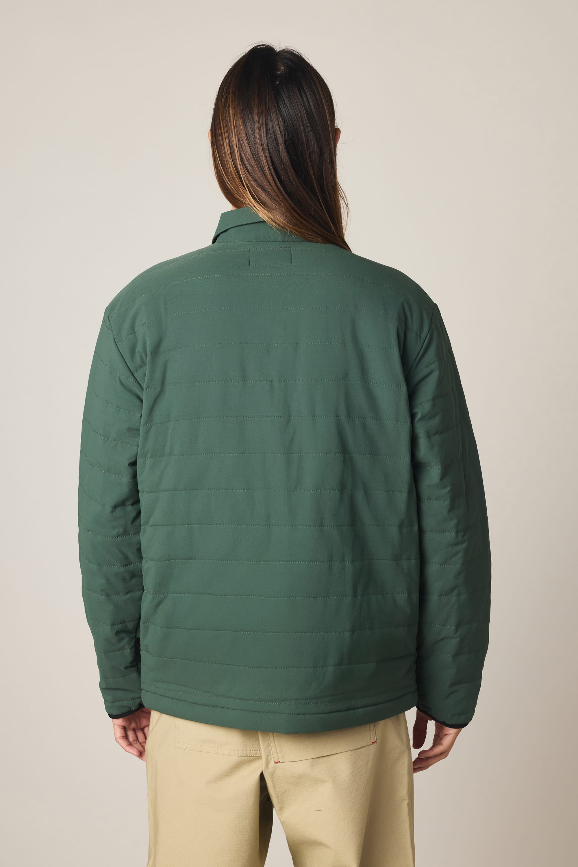 A person with long dark hair wears the 686 Men's Thermaldry Insulated Shacket, styled with beige pants, standing with their back to the camera against a plain, light background.
