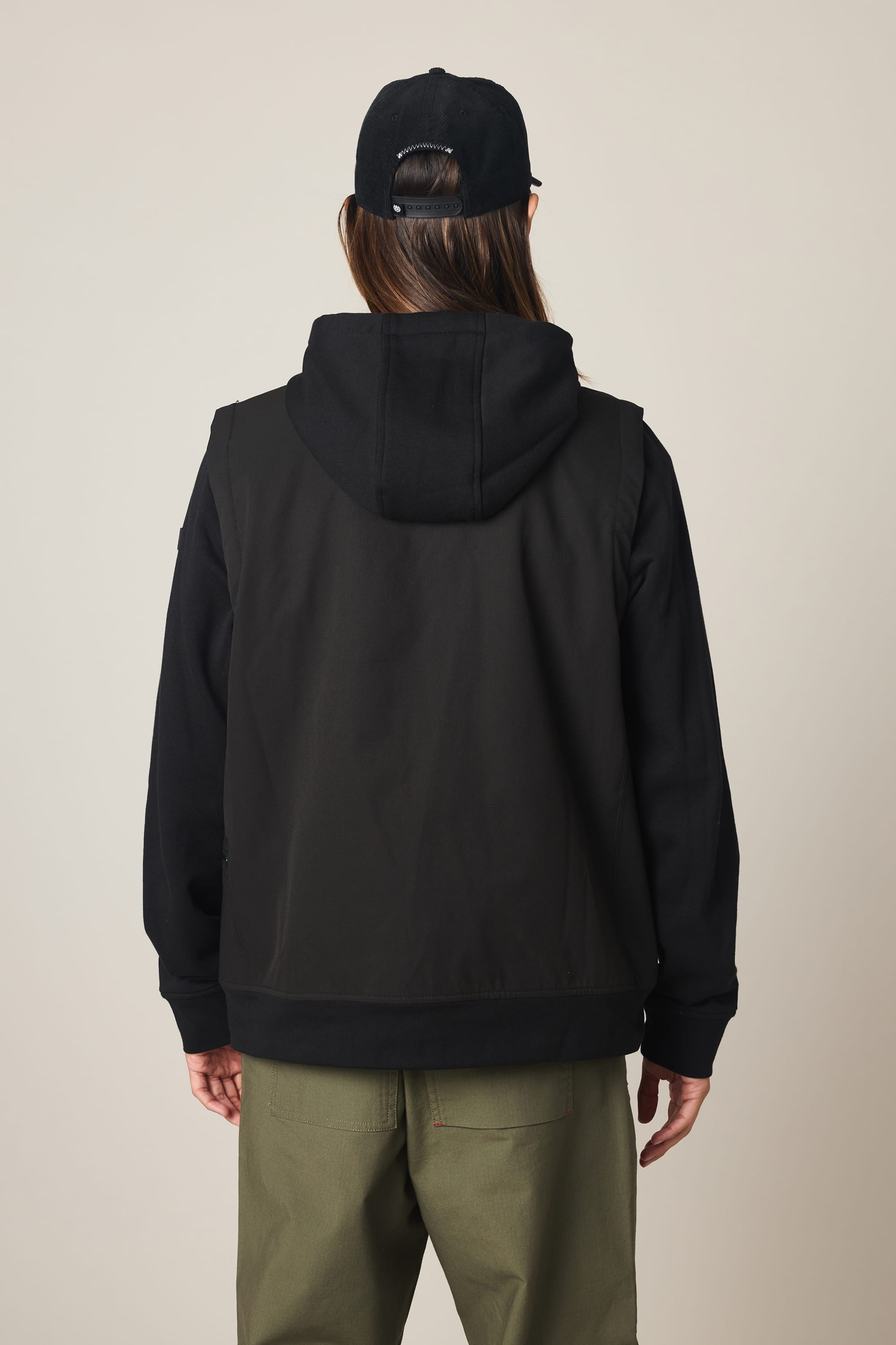 A person with long hair, wearing a black cap, the 686 Men's Merge Quilted Hoody by 686 layered as a vest, and olive green pants stands facing away from the camera against a light background.