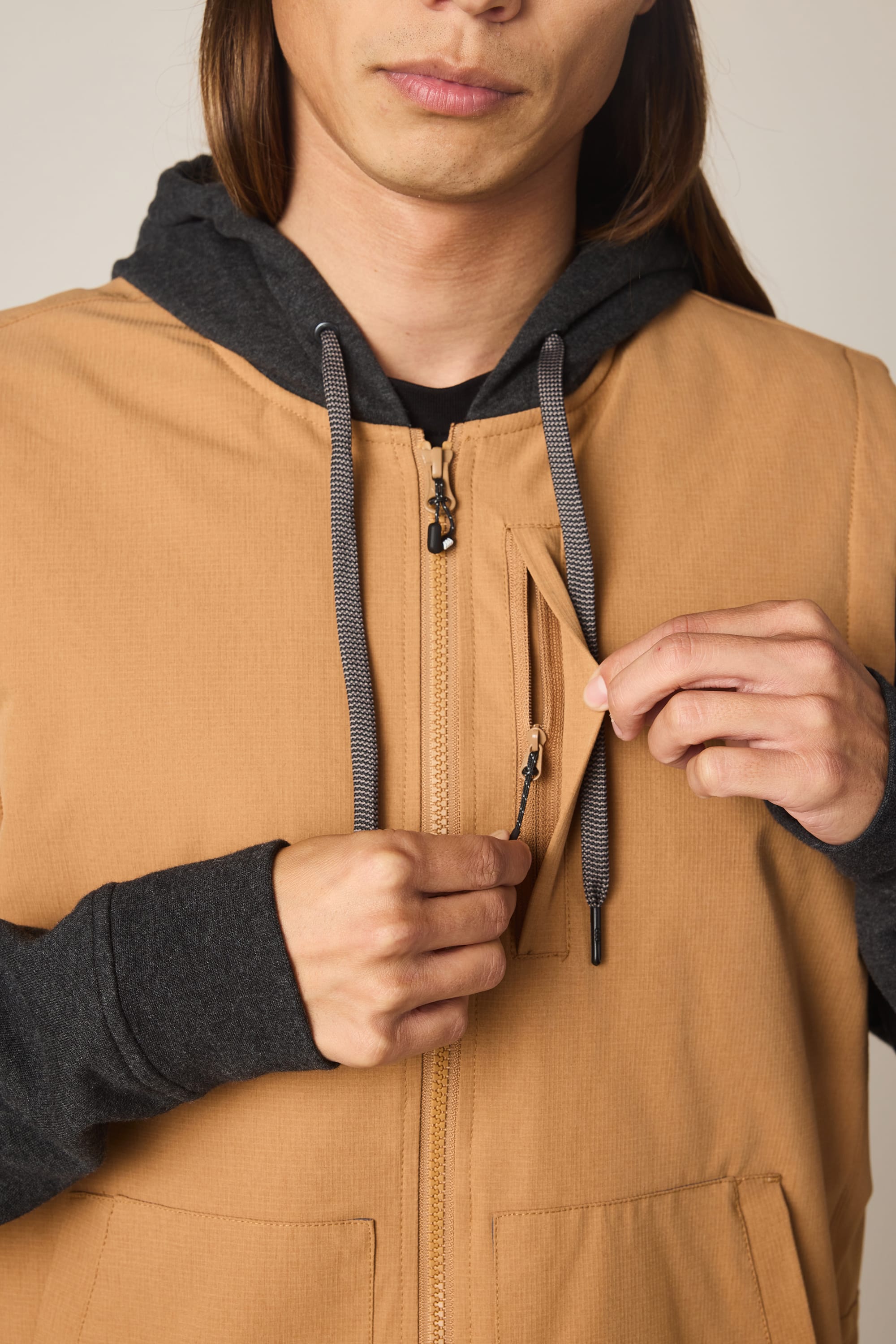 A person in the 686 Men's Merge Quilted Hoody, featuring a tan polyester shell and black hood, is unzipping a small chest pocket; only their lower face and torso are visible.