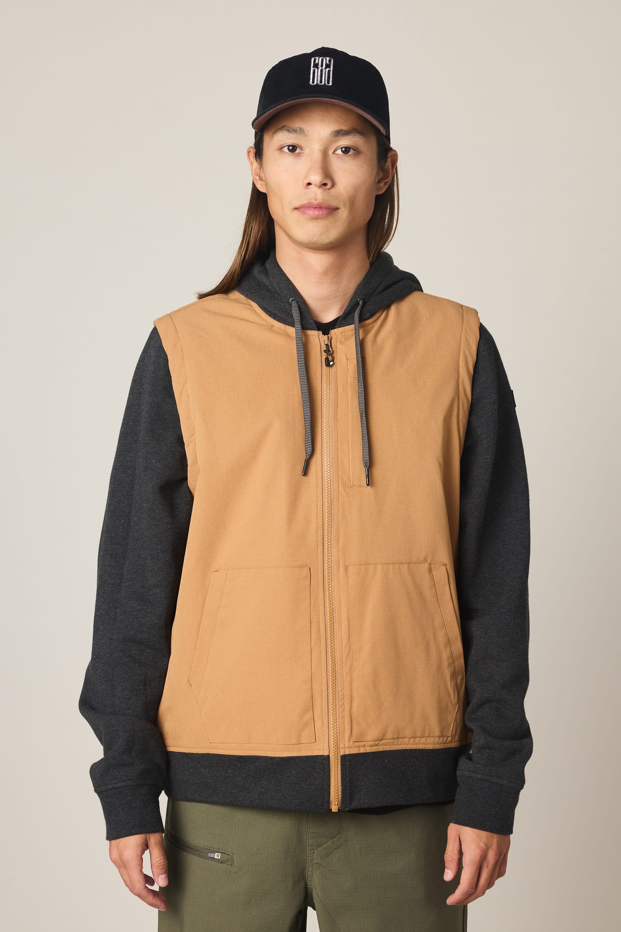 A person with long hair wears a black baseball cap and the 686 Men's Merge Quilted Hoody by 686, styled as a tan zip-up vest over a black hoodie, standing against a plain light background facing the camera.