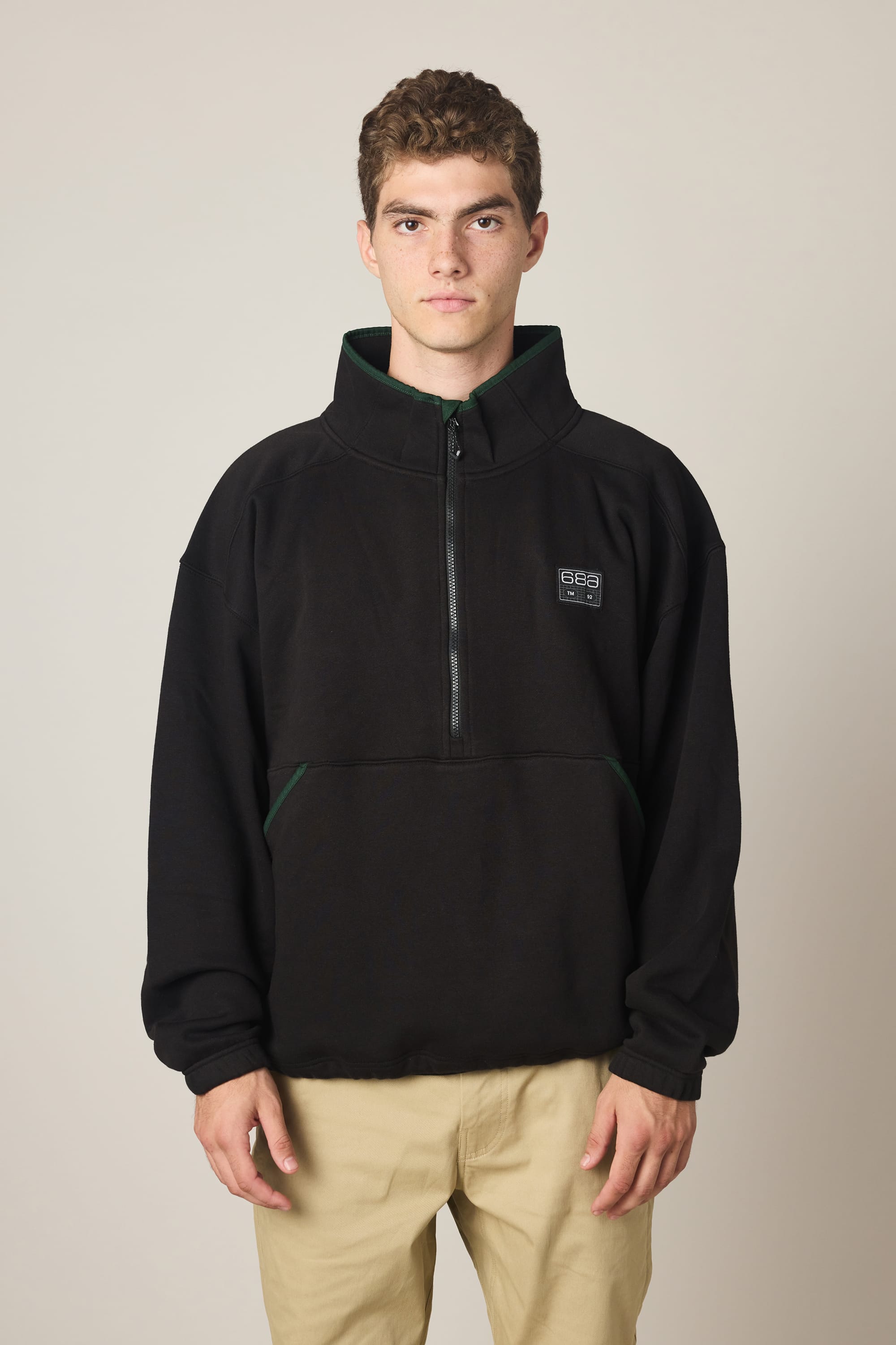 A young man with short curly hair wears the 686 Men's Burnout Anorak in black, paired with beige pants, standing facing forward against a plain light background.