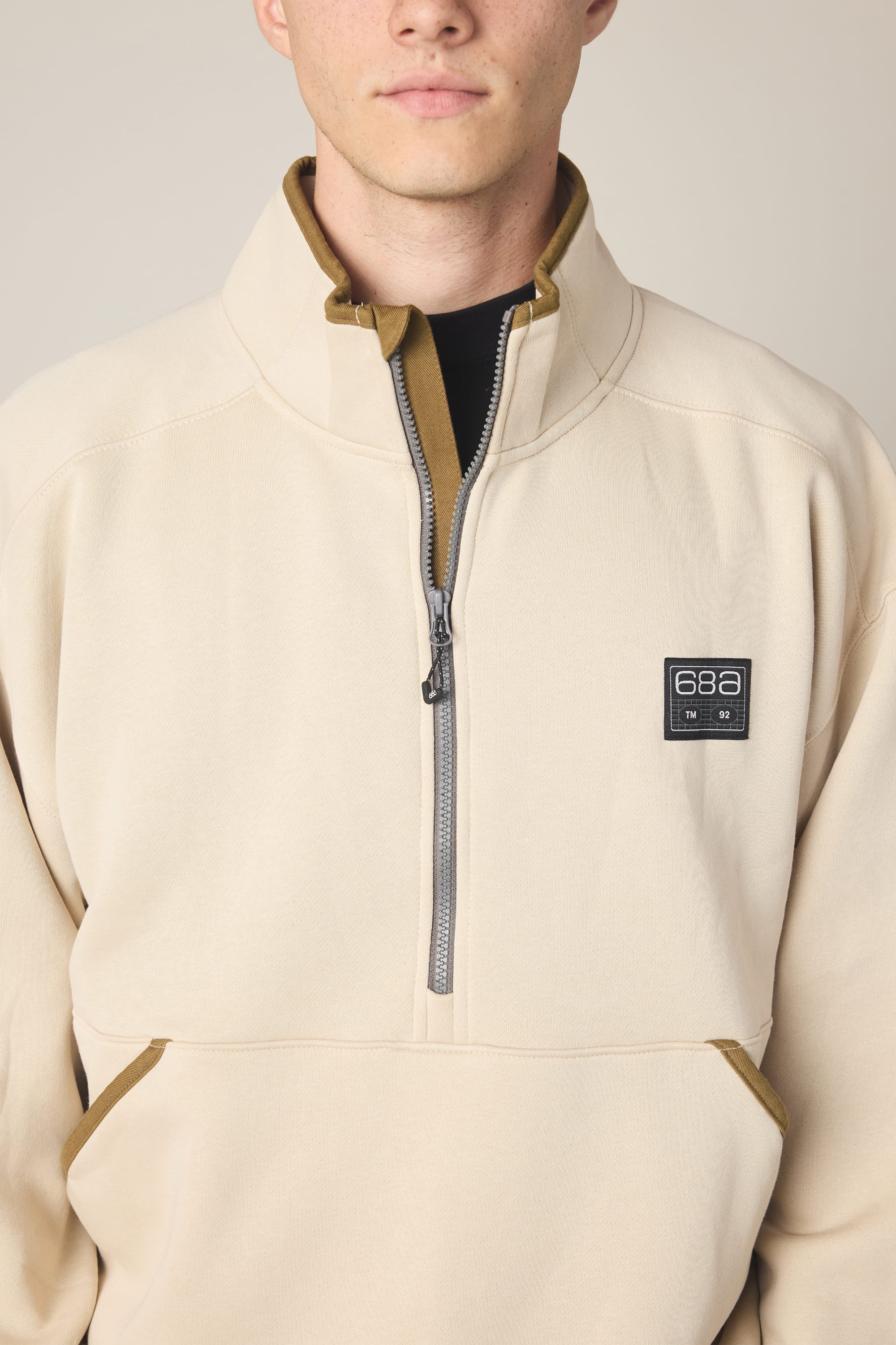 A person wears the 686 Men's Burnout Anorak, a light beige woven jacket with a high collar, quarter-zip style, black and white "686" chest patch, brown trim accents, and their face is partially visible.