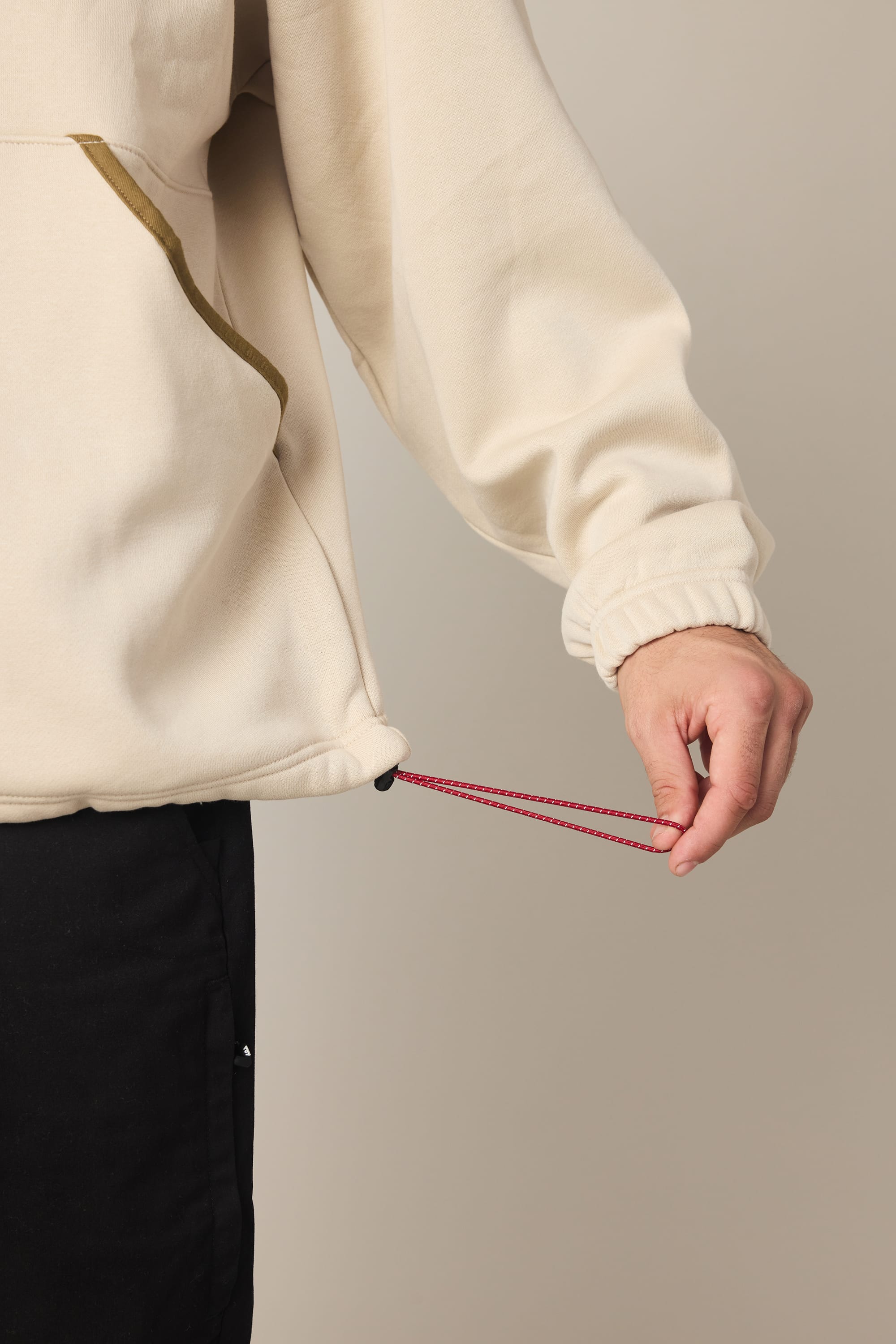 A man in the 686 Men's Burnout Anorak, a cream-colored woven jacket, adjusts the red drawstring at the hem while wearing black pants. The background is plain and neutral.