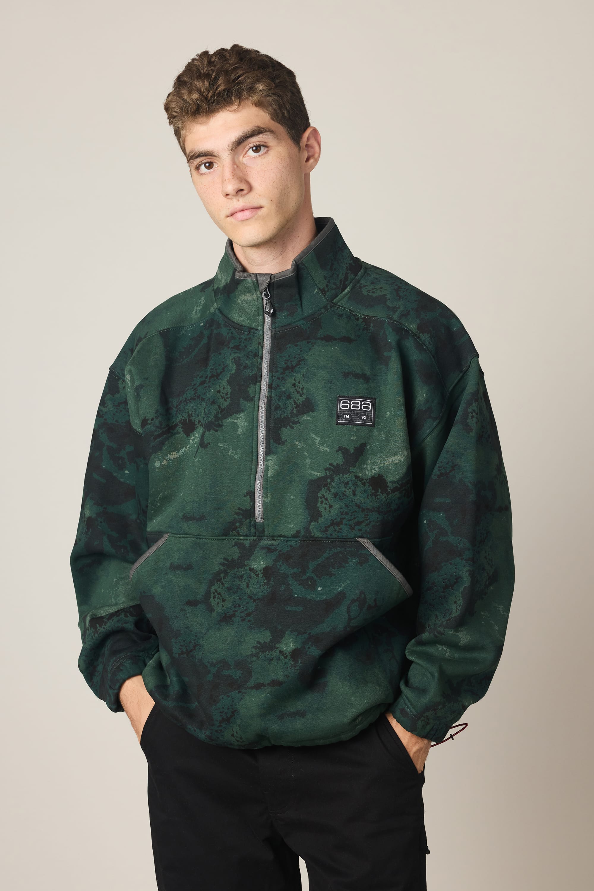 A young man with short curly hair wears the 686 Men's Burnout Anorak, a dark green and black half-zip woven jacket from 686, standing with his hands in his pockets against a plain light background.