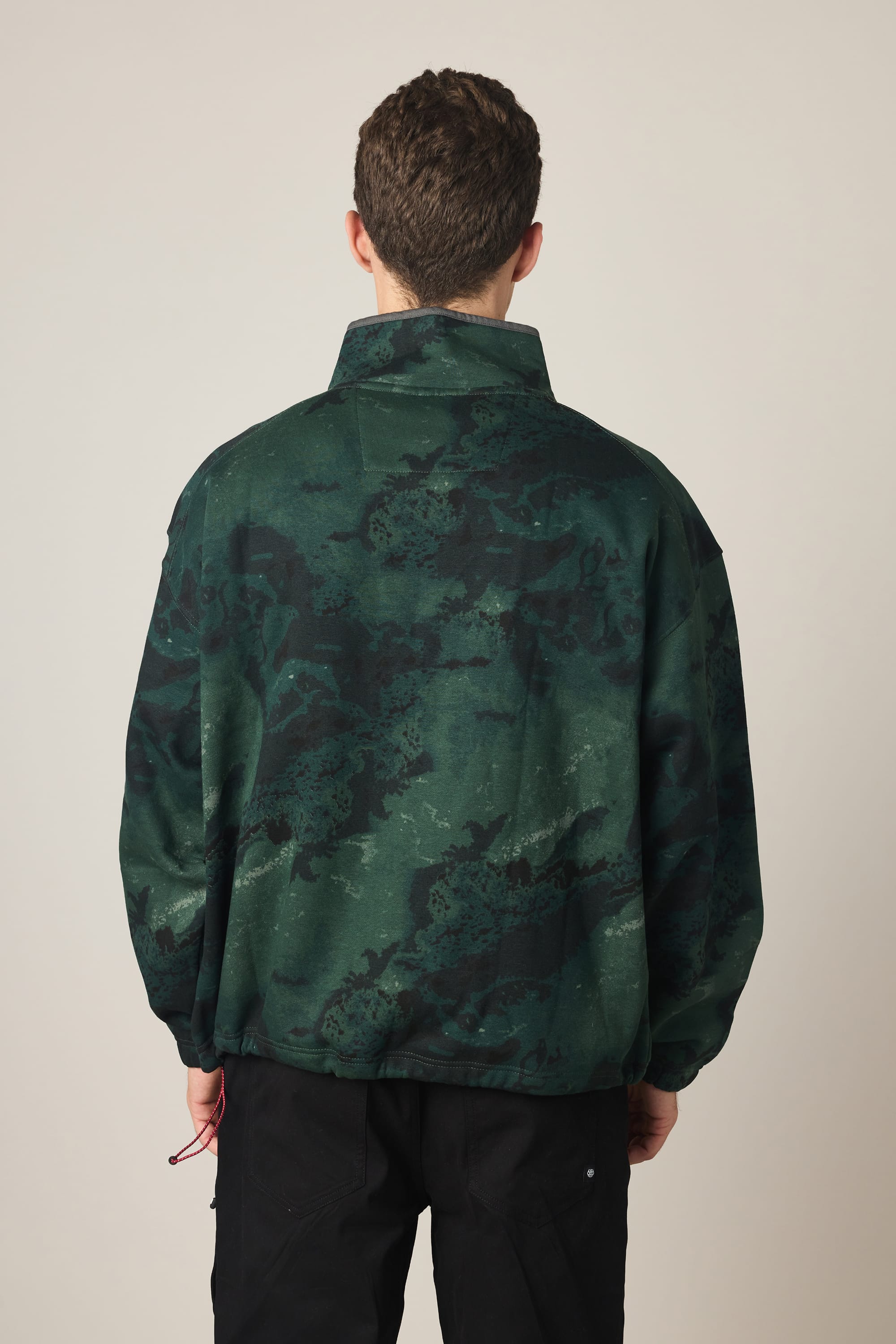 A person with short brown hair is shown from the back, wearing a 686 Men's Burnout Anorak by 686 in dark green and black, paired with black pants, standing against a plain light background.