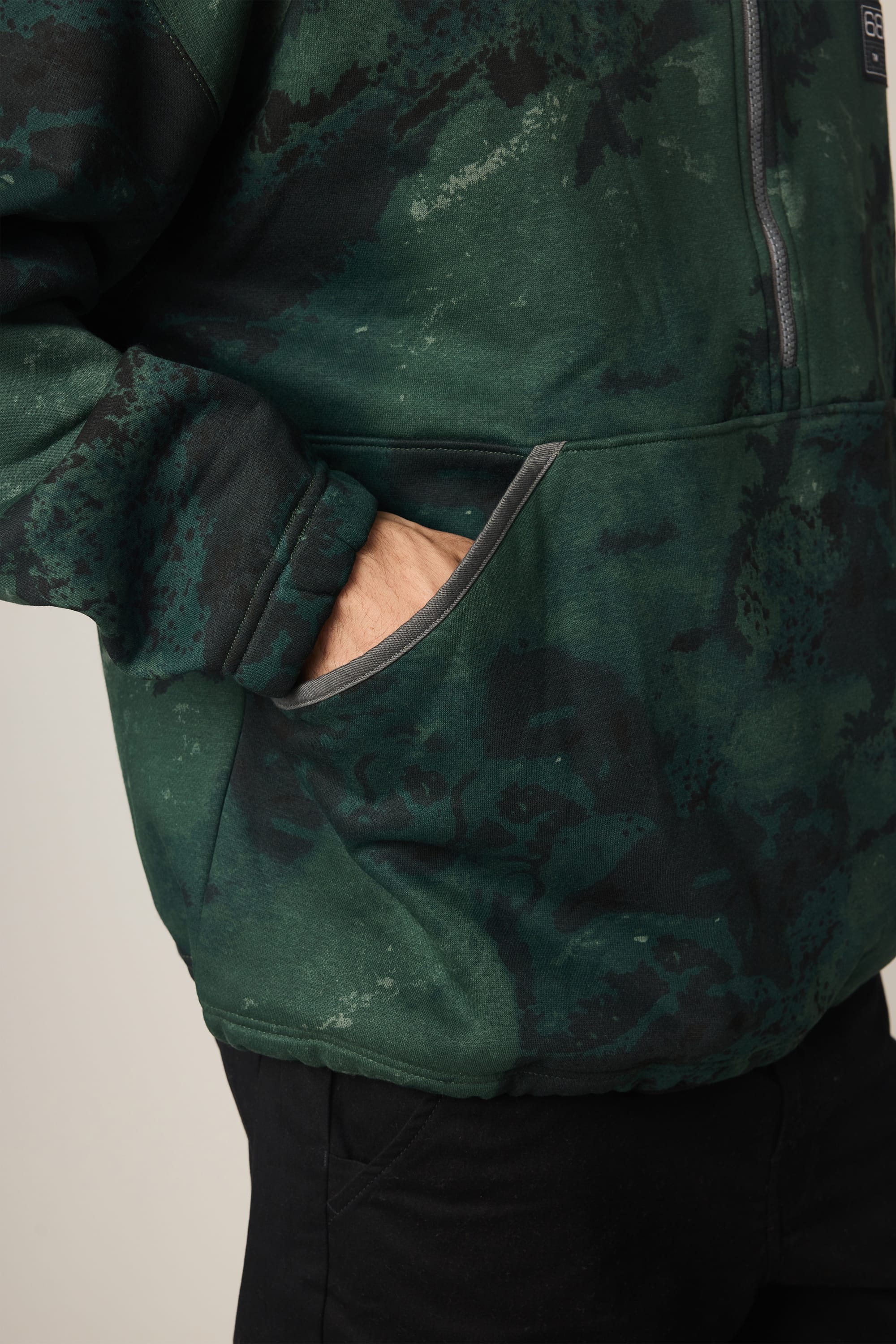 A person wears the 686 Men's Burnout Anorak, a green and black tie-dye hoodie by 686, with one hand in the front pocket; shown from the waist up, cropped at the shoulder, styled like a men's waterproof jacket.