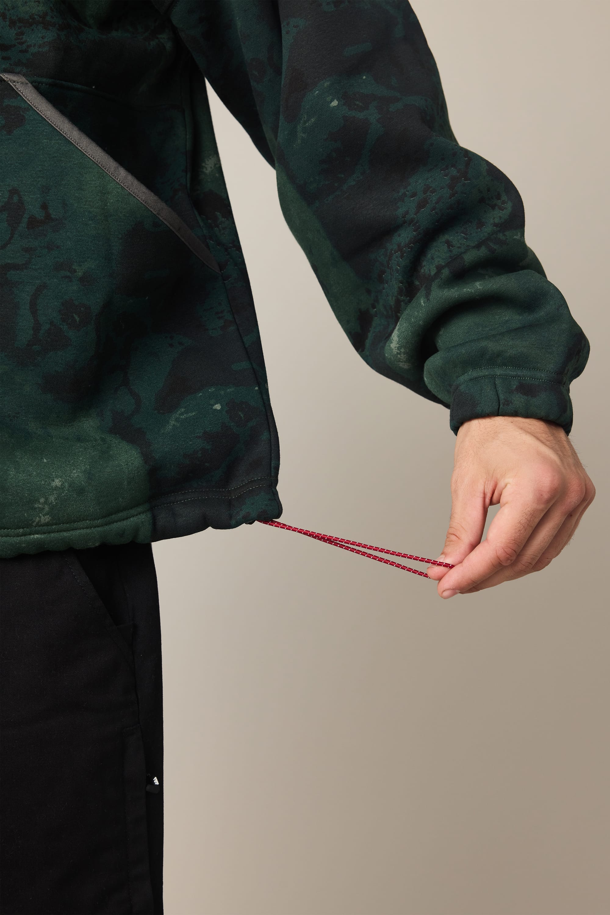 A person adjusts the red drawstring at the hem of a 686 Men's Burnout Anorak jacket in dark green and black, paired with black pants. Only their hand and part of the torso are visible.