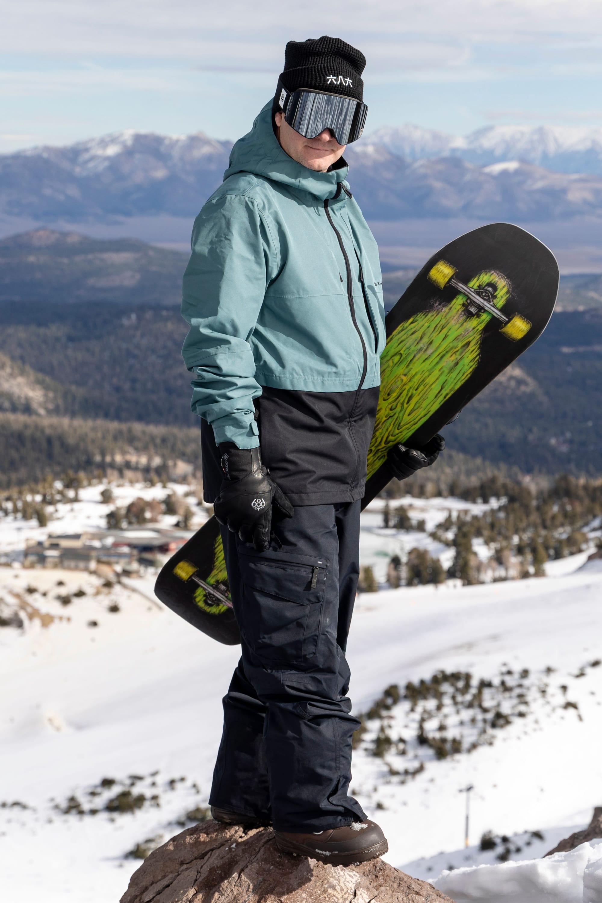 A person dressed in 686 Men’s SMARTY® 3-in-1 Cargo Pant stands on a snowy mountain holding a snowboard with a green graphic, with snow-covered landscapes and mountains visible in the background.