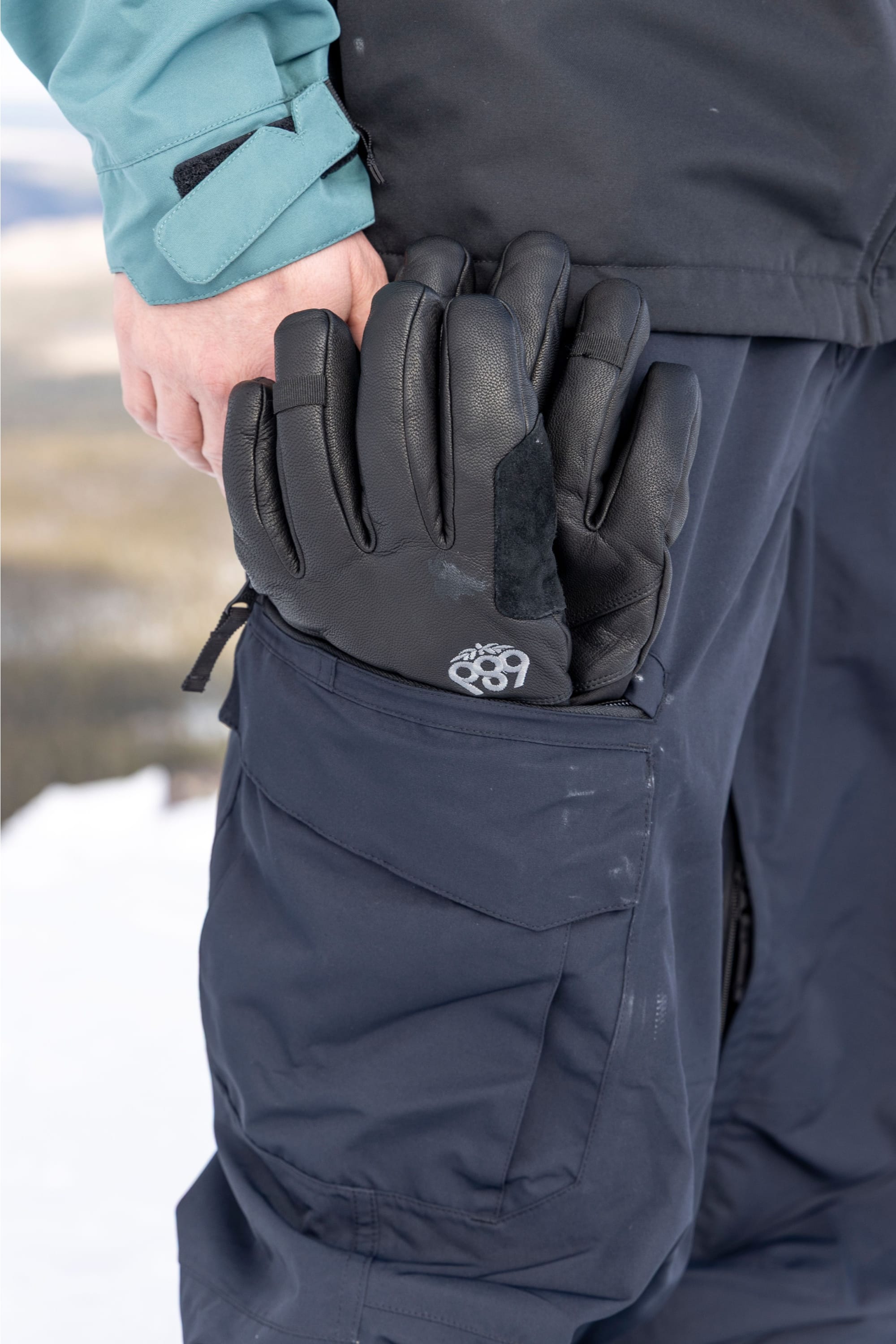 A person dressed in winter attire stands outdoors in the snow, holding black gloves partially tucked into the pocket of 686 Men’s SMARTY® 3-in-1 Cargo Pants by 686.