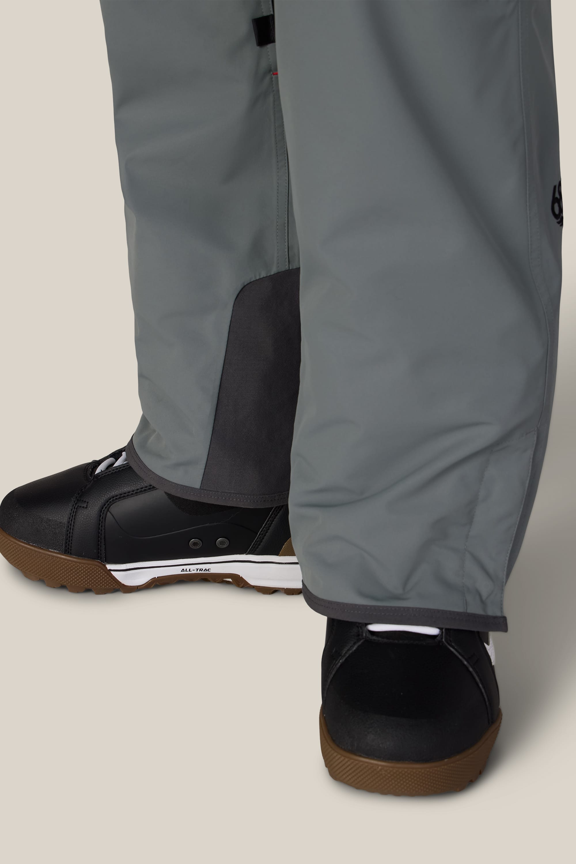 Close-up of a person wearing the 686 Men’s SMARTY® 3-in-1 Cargo Pant in grey and black athletic shoes with brown soles, standing on a light beige surface.