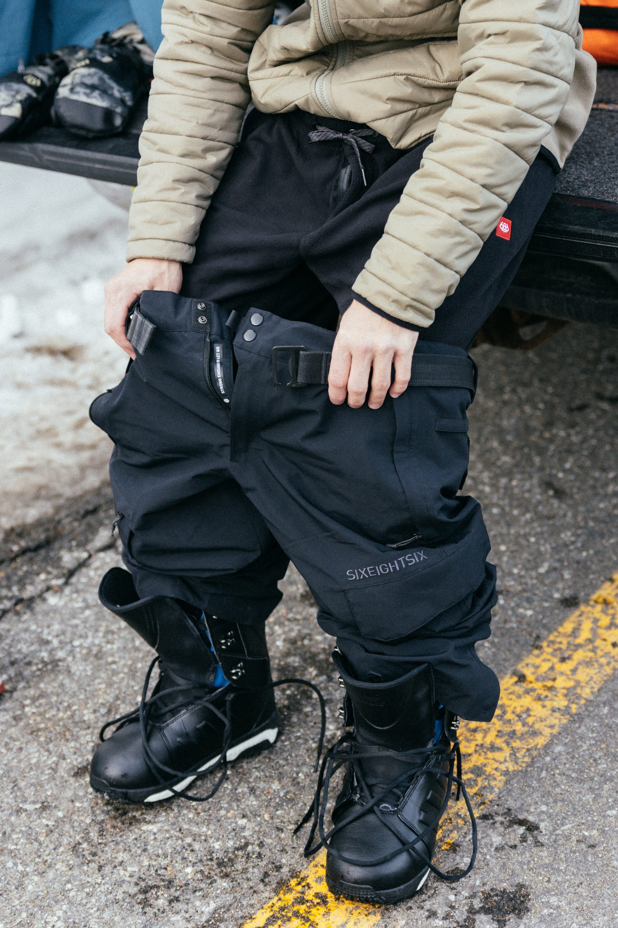 A person sitting on a car trunk puts on 686 Men’s SMARTY® 3-in-1 Cargo Pants and black snow boots. The ground is wet and snowy, and the person’s tan jacket sleeves are visible.