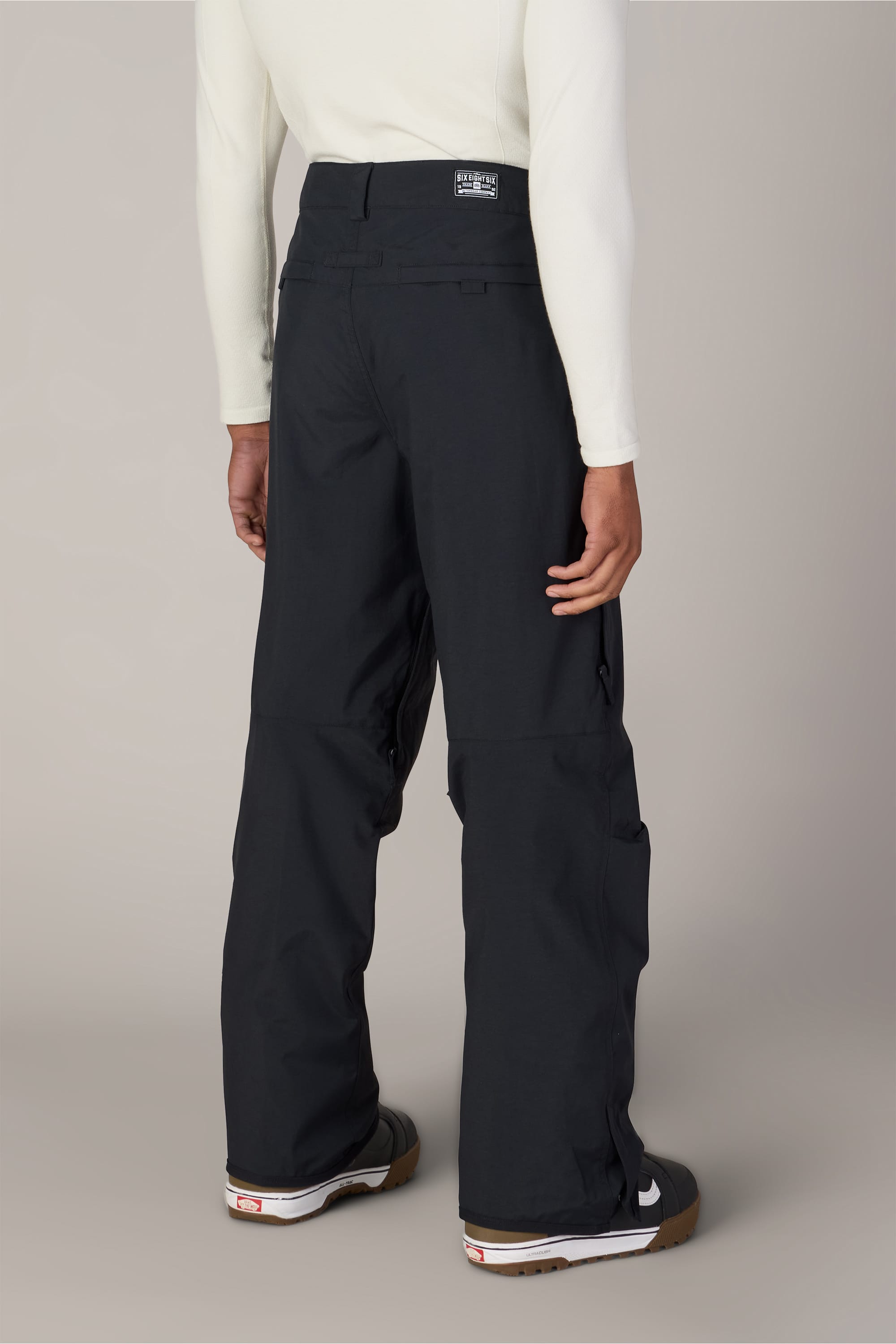 A person is standing with their back facing the camera, wearing a long-sleeve white shirt and the 686 Men’s Standard Shell Pant by 686—affordable, loose-fitting black pants—paired with black shoes featuring white soles. The background is plain and neutral.