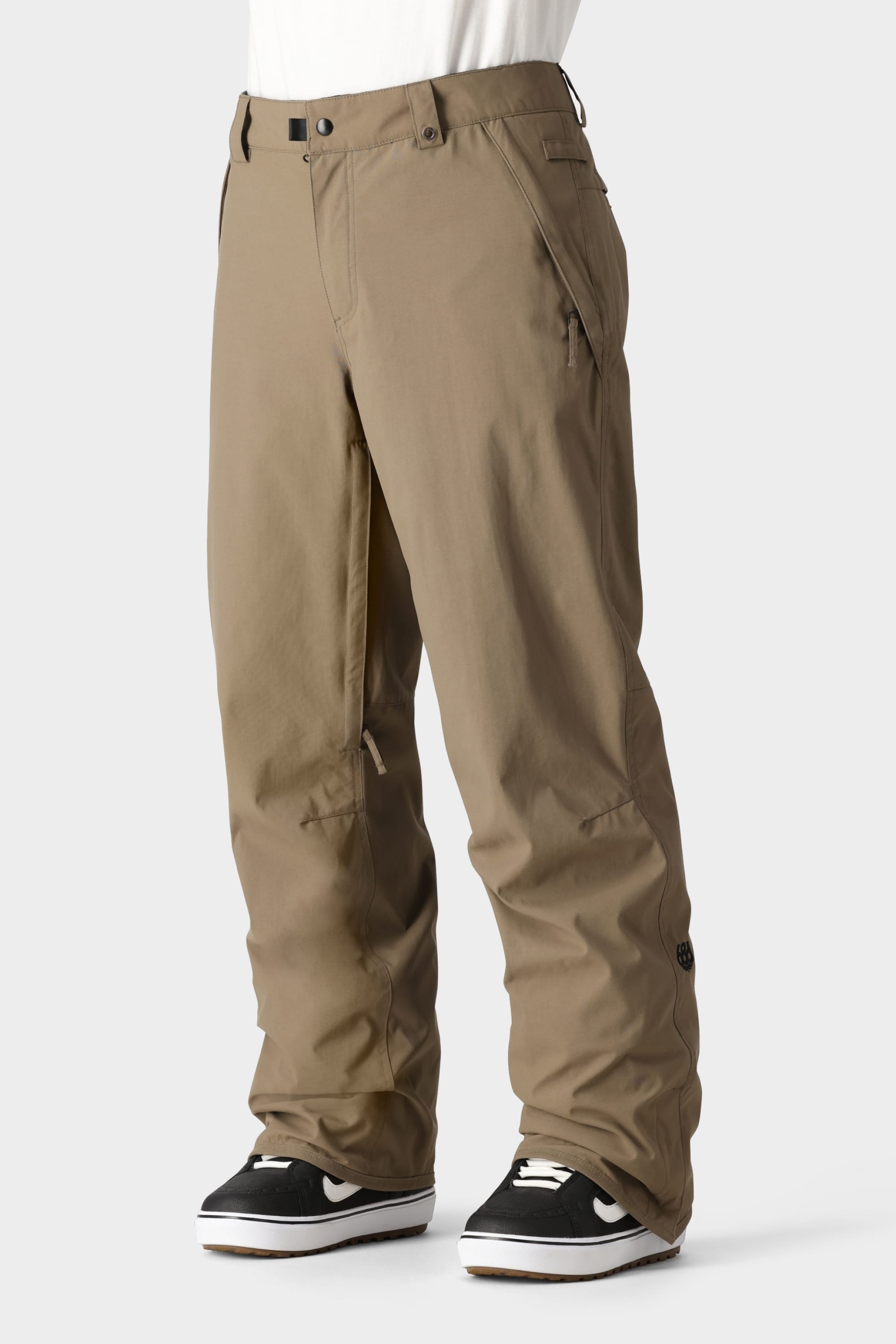 A person wearing the 686 Men’s Standard Shell Pant in a loose-fitting, affordable light brown style, paired with black-and-white sneakers, shown from the waist down against a plain light background.