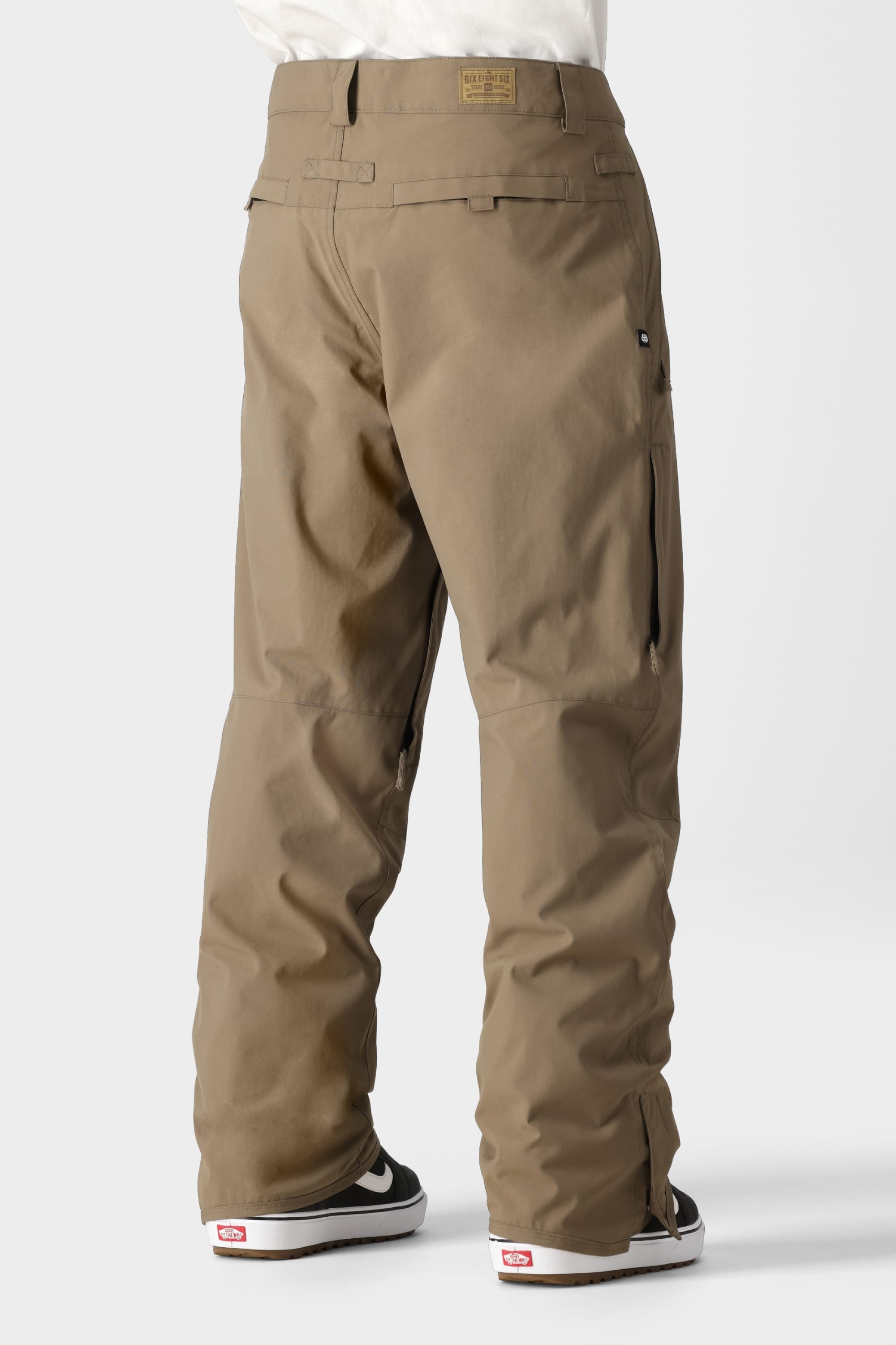 A person wearing the 686 Men’s Standard Shell Pant by 686—loose-fitting, affordable khaki pants with pockets—paired with black-and-white Vans sneakers, stands against a plain light background. The back view highlights seam and waistband details on the pants.