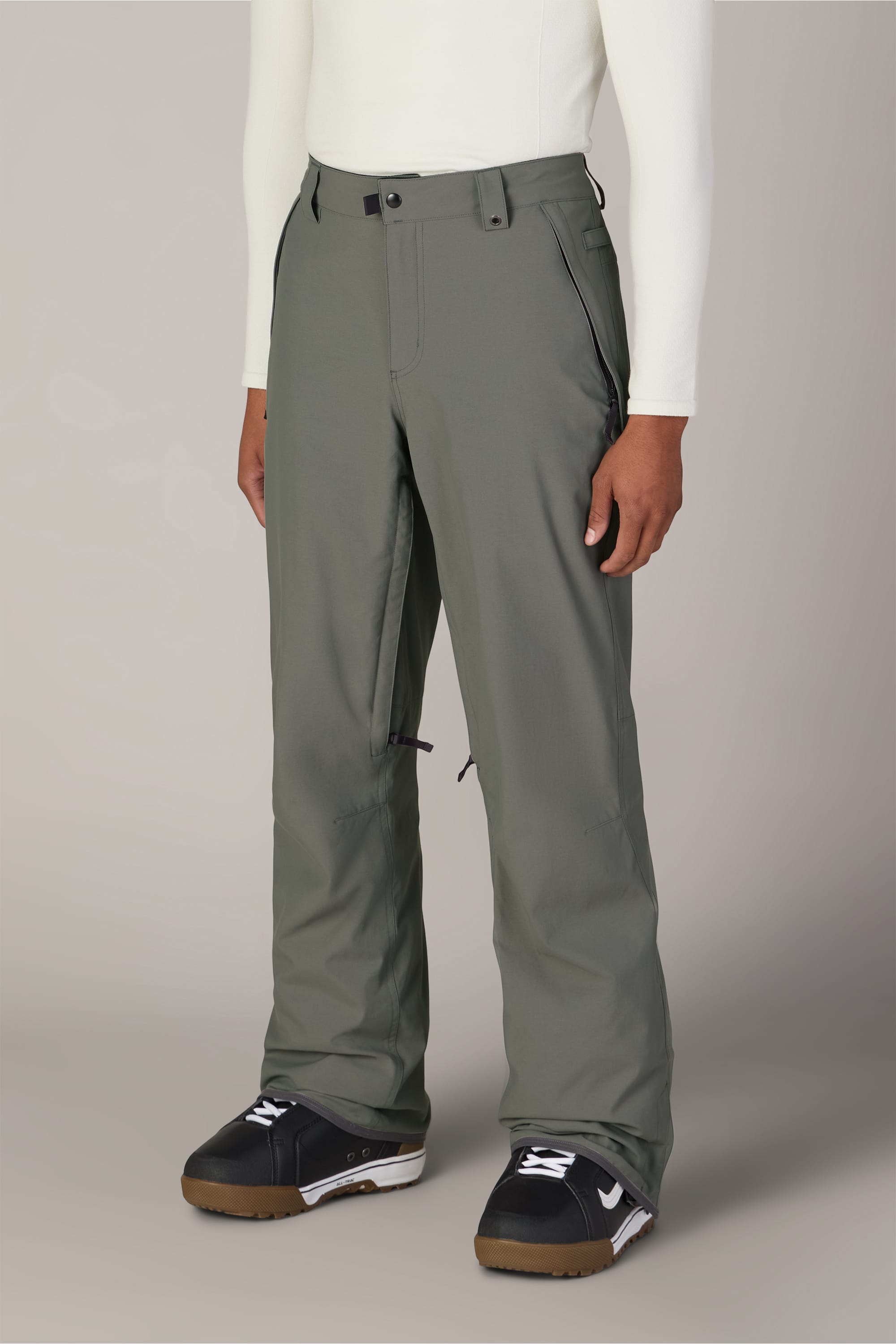 A person wearing 686 Men’s Standard Shell Pant in sage green, a white long-sleeve top, and black and white snow boots stands against a plain light gray background.