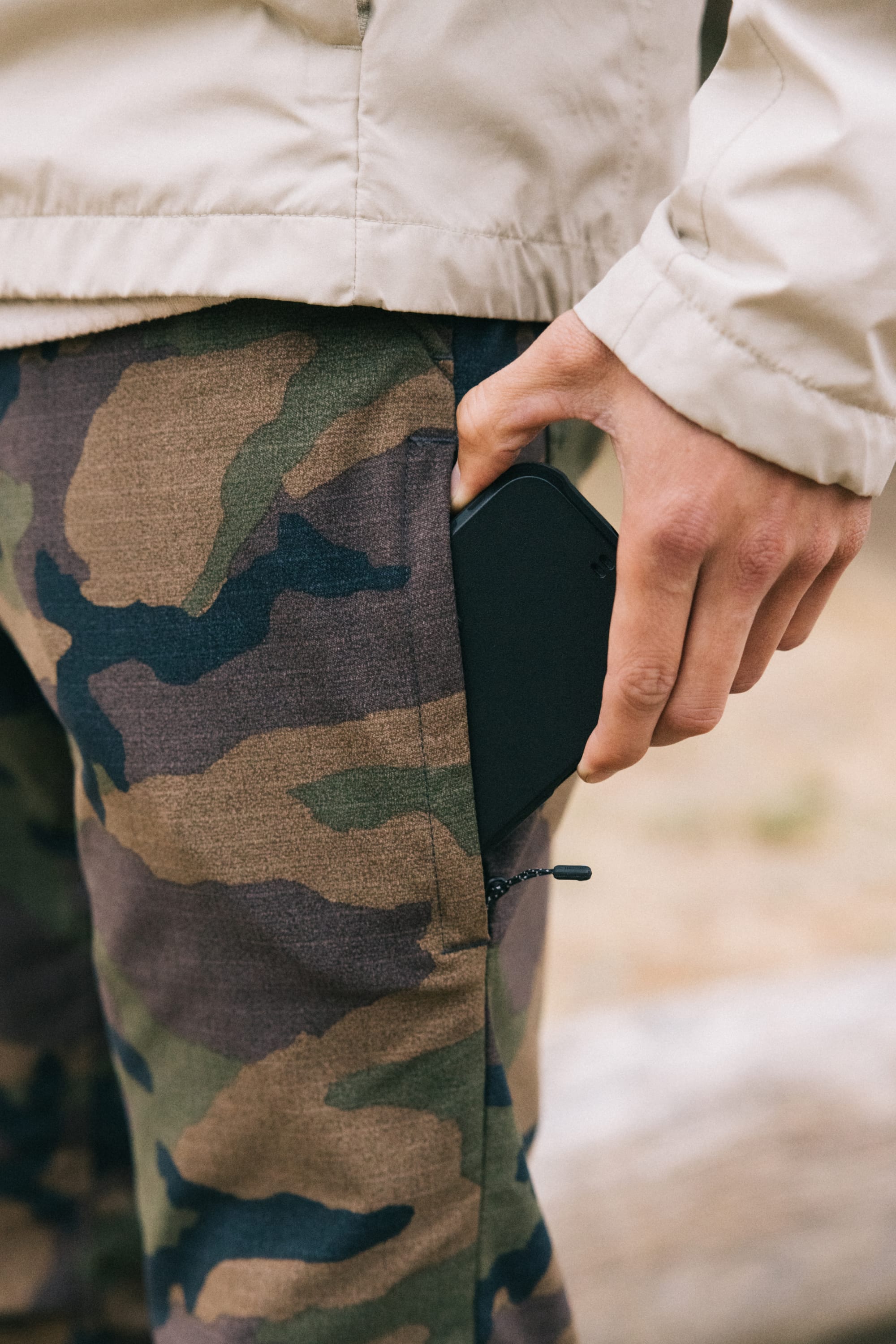 A person wearing 686 Men's Everywhere® Pant - Slim Fit in camouflage and a beige jacket puts a black smartphone into their pocket.