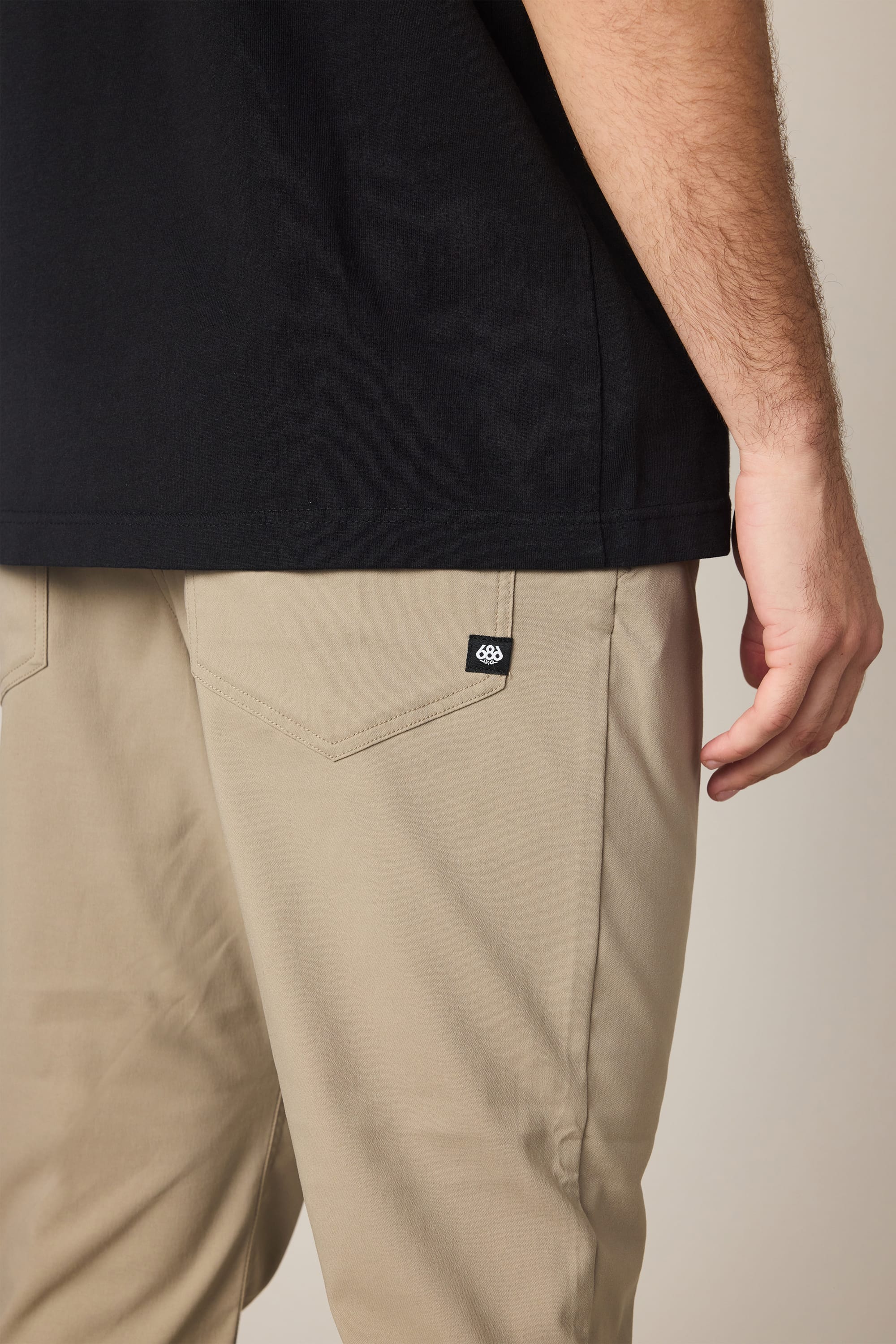 A person wearing 686 Men's Everywhere® Pant - Slim Fit in beige and a black shirt stands with their right arm relaxed by their side. A small black tag featuring the 686 brand is visible on the back pocket of the breathable pants.