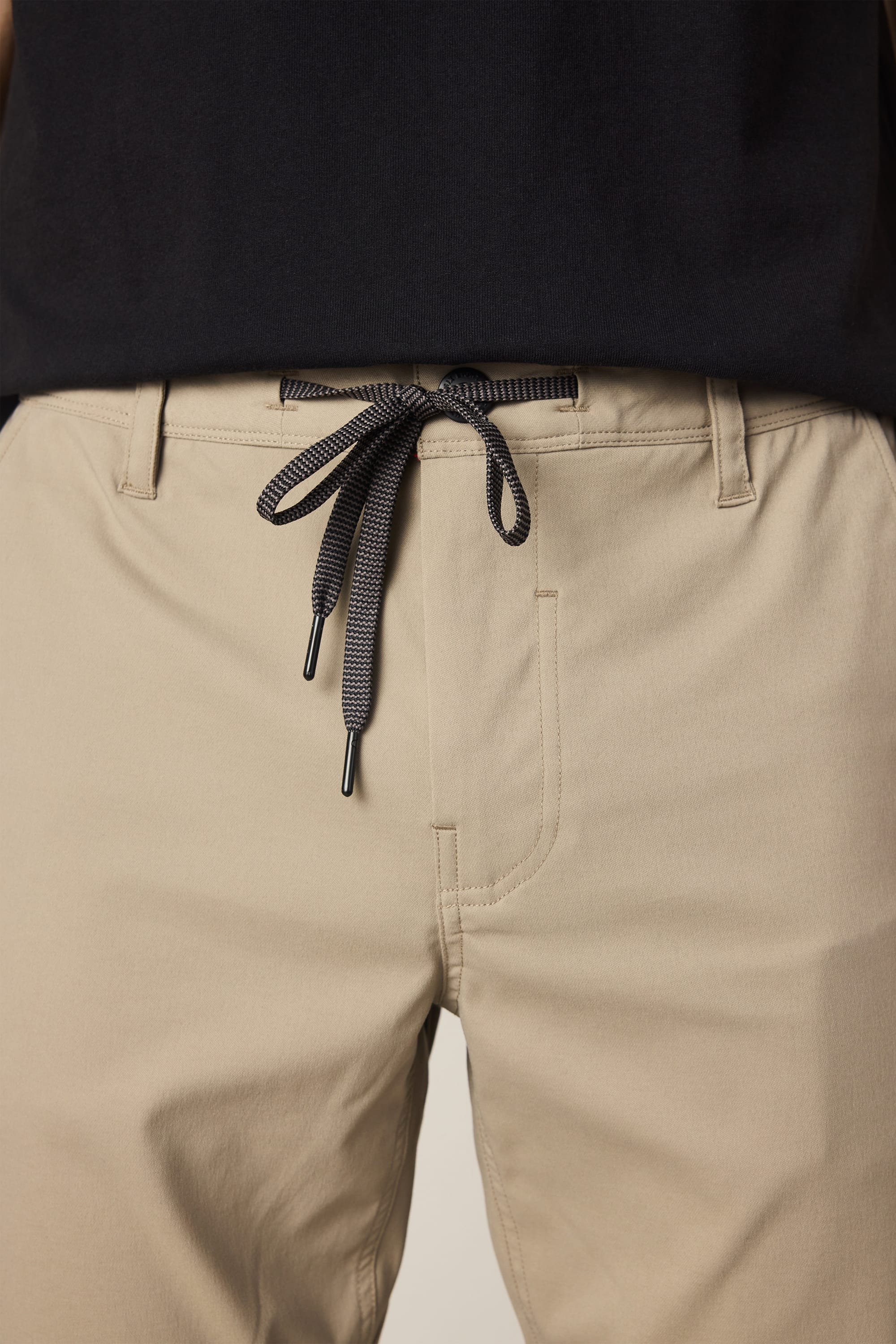 Close-up of a person wearing the 686 Men's Everywhere® Pant - Slim Fit in beige, featuring a black drawstring tie at the waist paired with a black shirt. The image focuses on the waist and upper thighs, highlighting the bow-tied drawstring and showcasing these stain-resistant, breathable pants by 686.