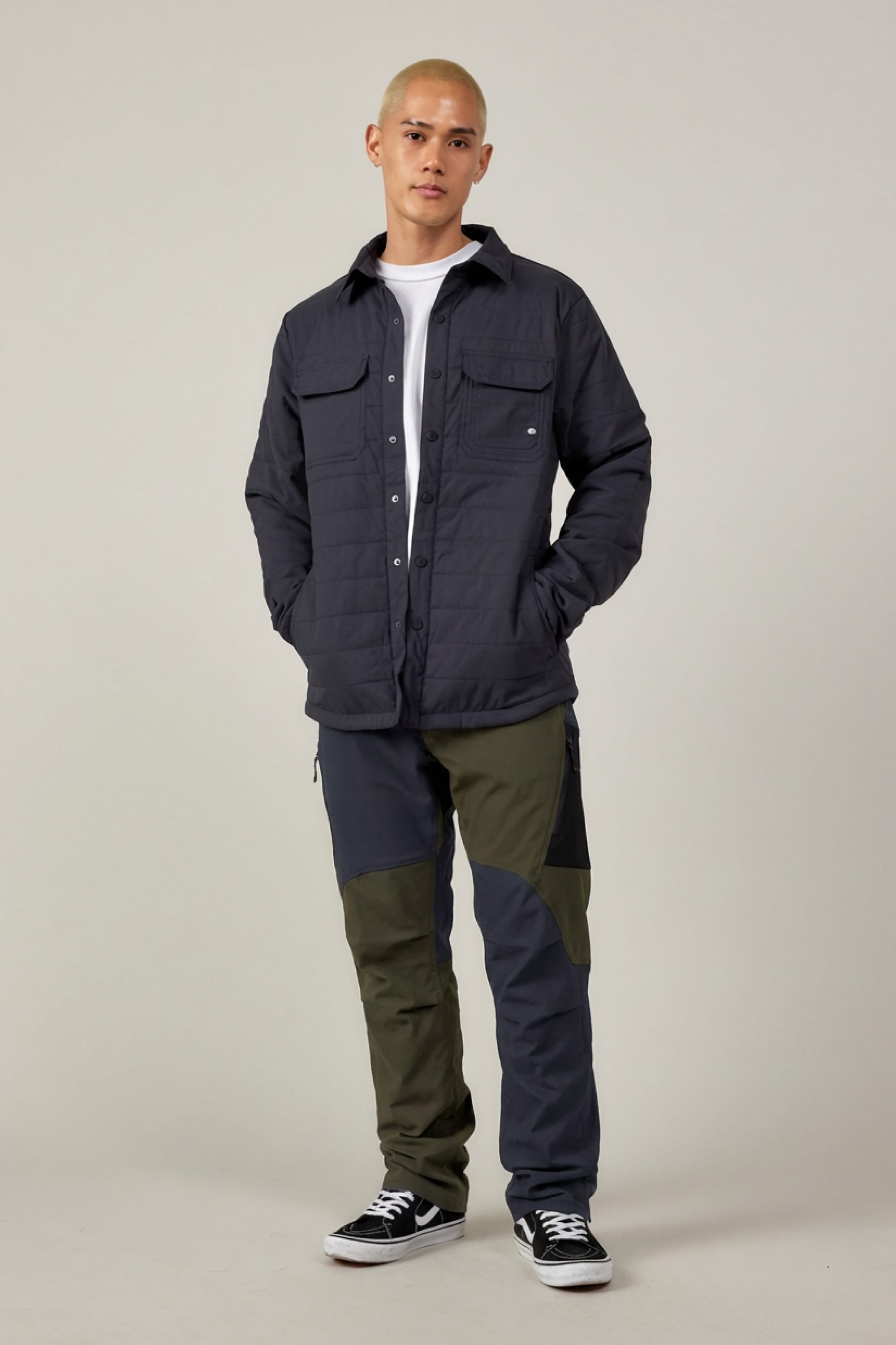 A person with short blond hair faces the camera, wearing a dark quilted jacket over a white shirt, 686 Men's Anything Cargo Pant - Relaxed Fit in two-tone dark and olive, and black sneakers with white soles on a plain background.