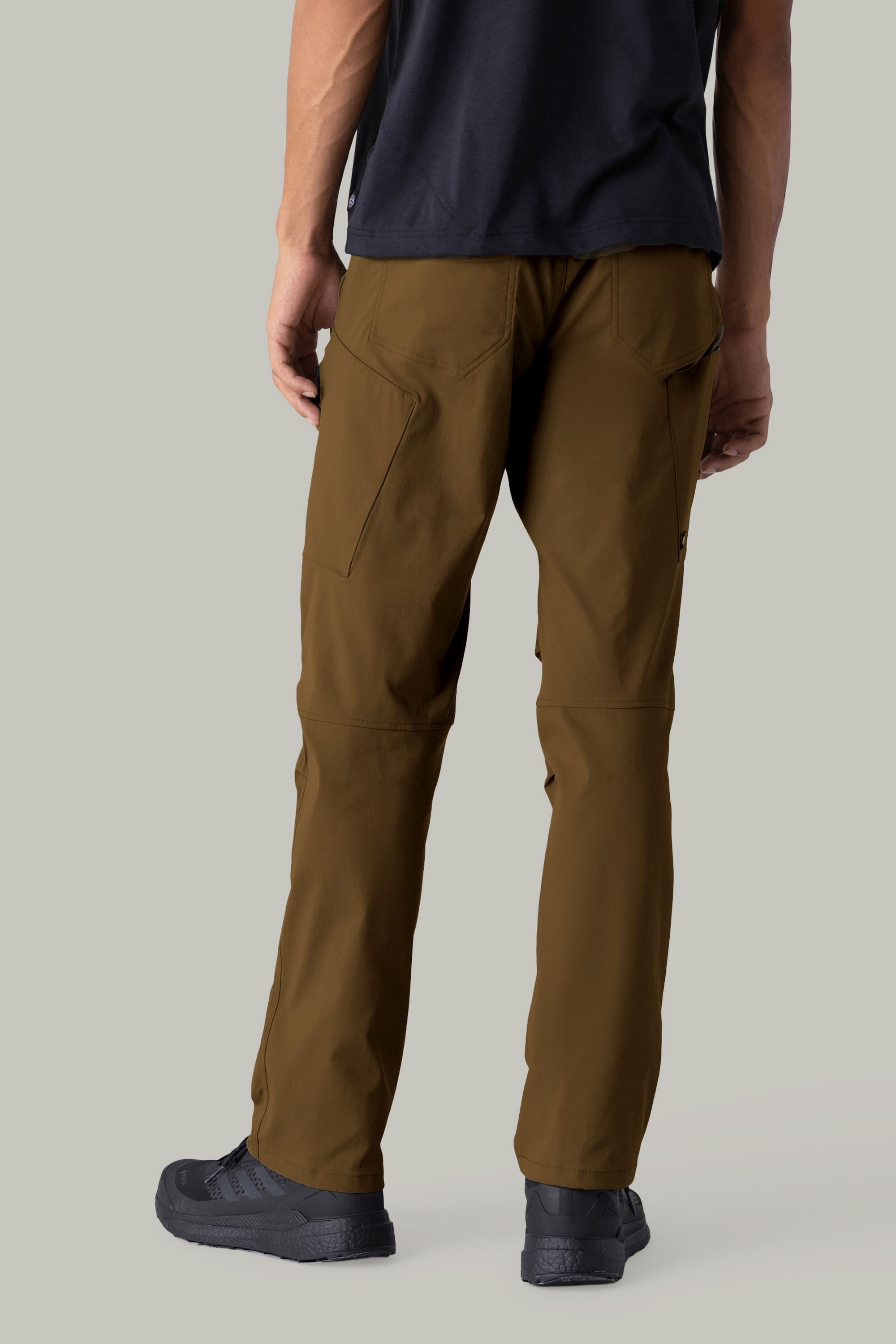 A person wearing 686 Men's Anything Cargo Pant - Relaxed Fit in brown, a black T-shirt, and black sneakers stands facing away against a plain light gray background.